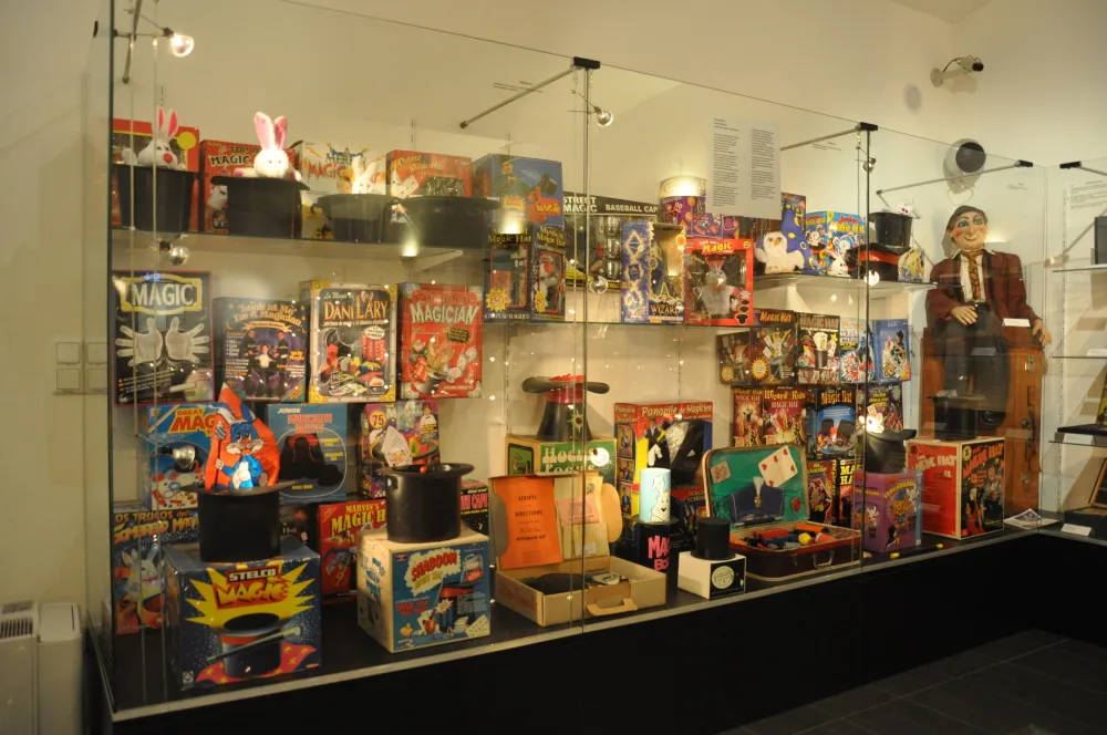 Glass display case filled with colorful magic trick boxes, props, and a mannequin in a suit