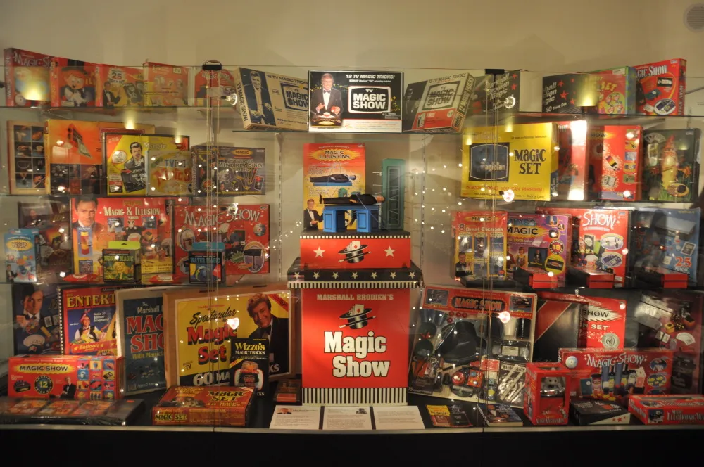 Display case filled with vintage magic trick boxes and kits, including a red 'Magic Show' box in the center and various colorful packages on shelves