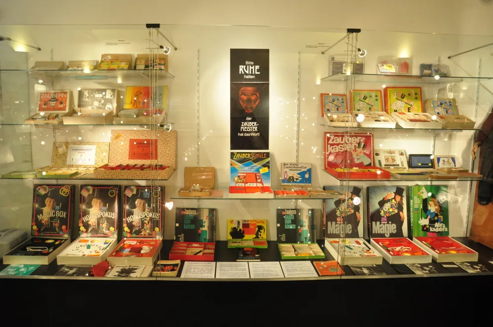 glass display case filled with magic books, trick boxes, and memorabilia, with a poster in the center