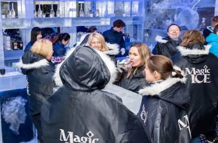 Grupa odwiedzających w czarnych pelerynach termicznych z białym futrzanym wykończeniem i logo Magic Ice, spędzających czas w lodowym barze, w tle widoczne lodowe rzeźby i lada barowa.