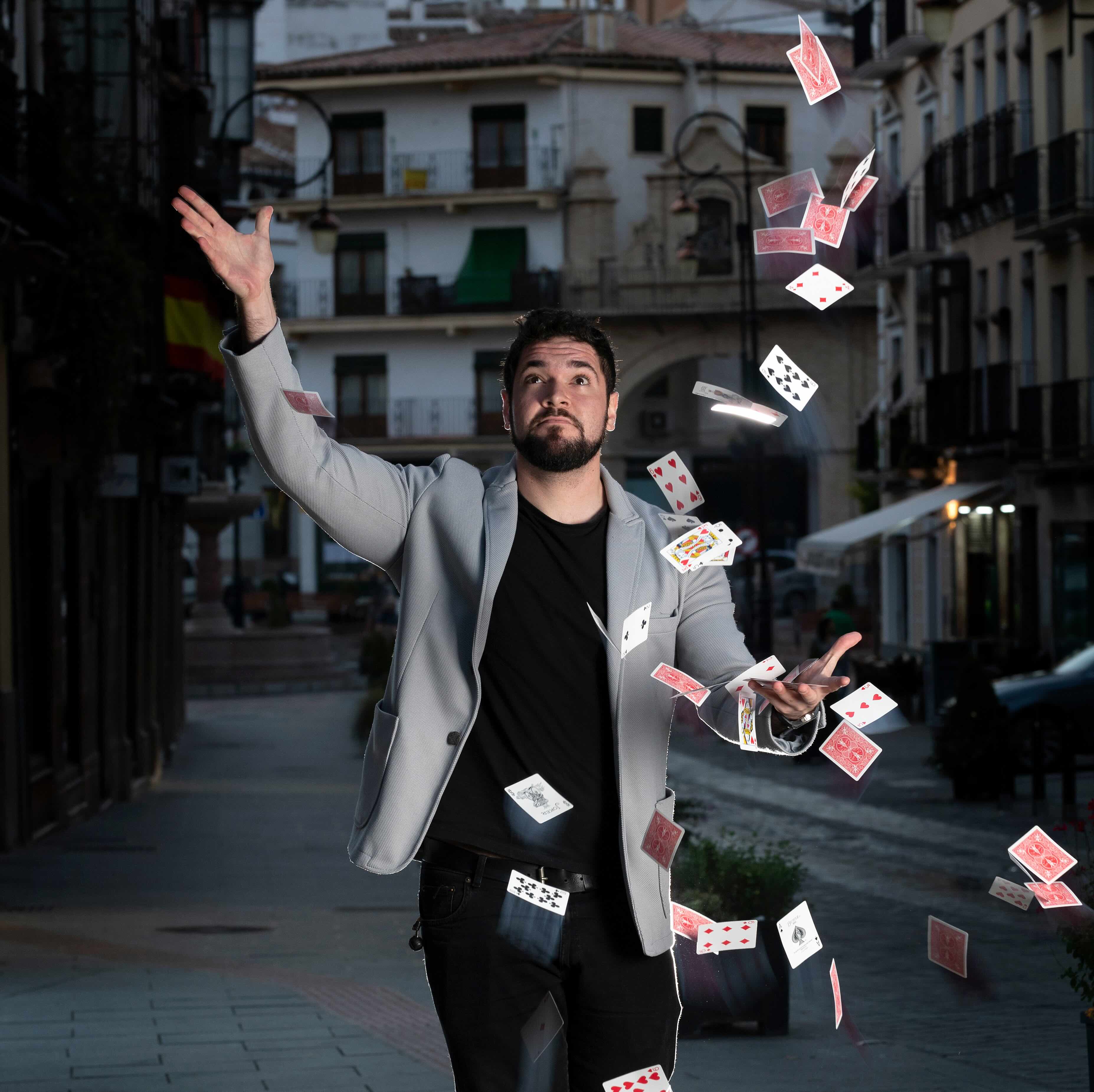 A man in a gray blazer and black shirt performs a card trick on a city street, with playing cards floating around him.