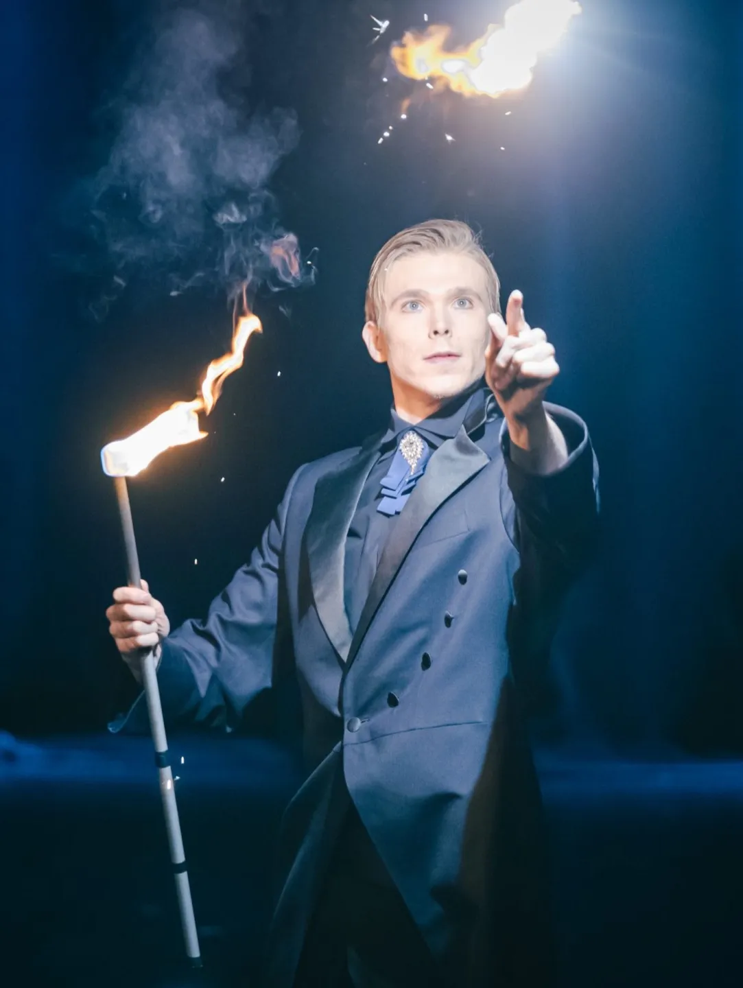 Magik w ciemnym garniturze trzymający płonącą pochodnię na scenie z iskrami i dymem