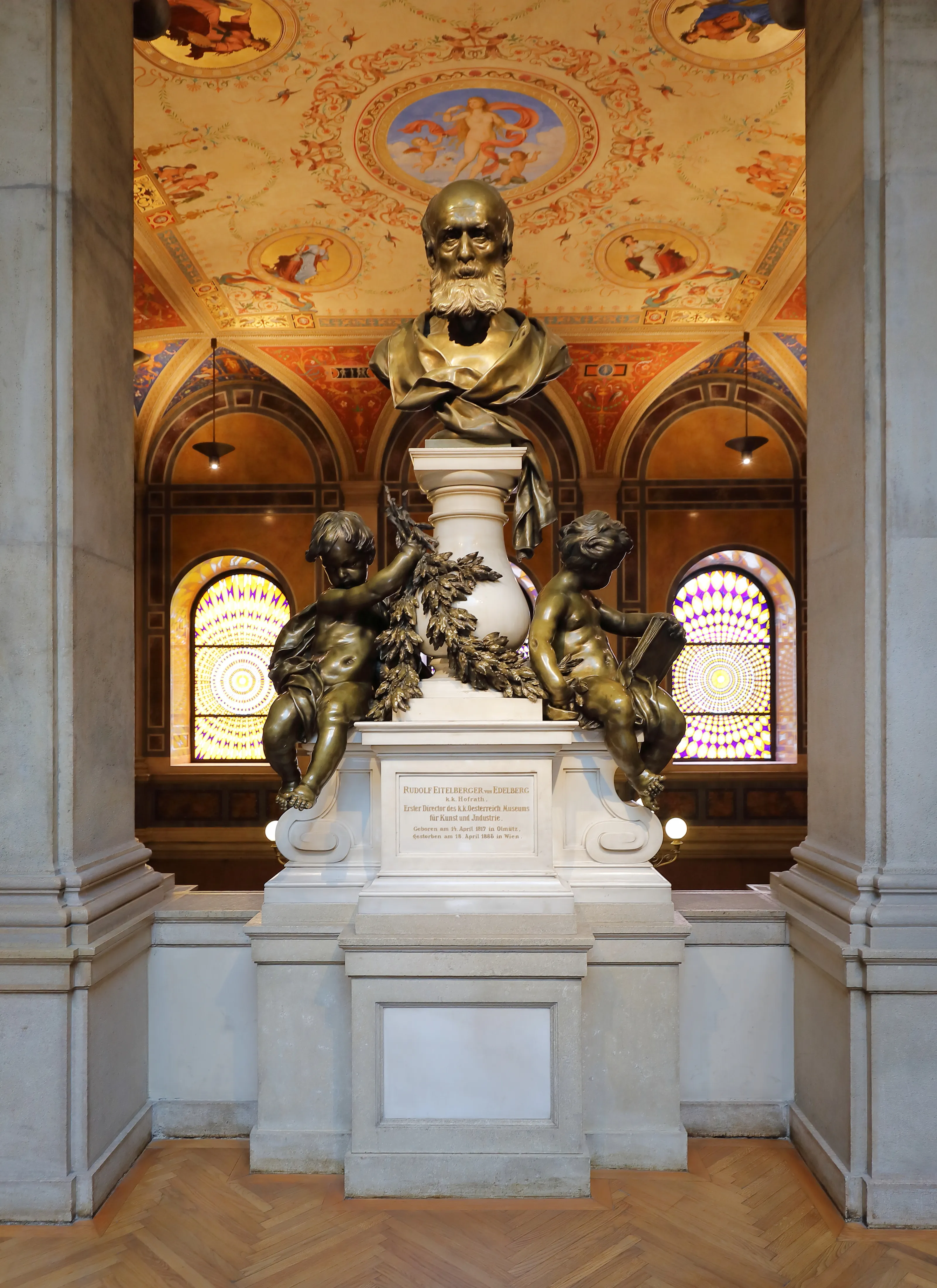 Bronzen buste van een bebaarde man op een wit marmeren sokkel, geflankeerd door twee cherubijnse figuren, in een grote zaal met sierlijke plafondfresco's en glas-in-loodramen