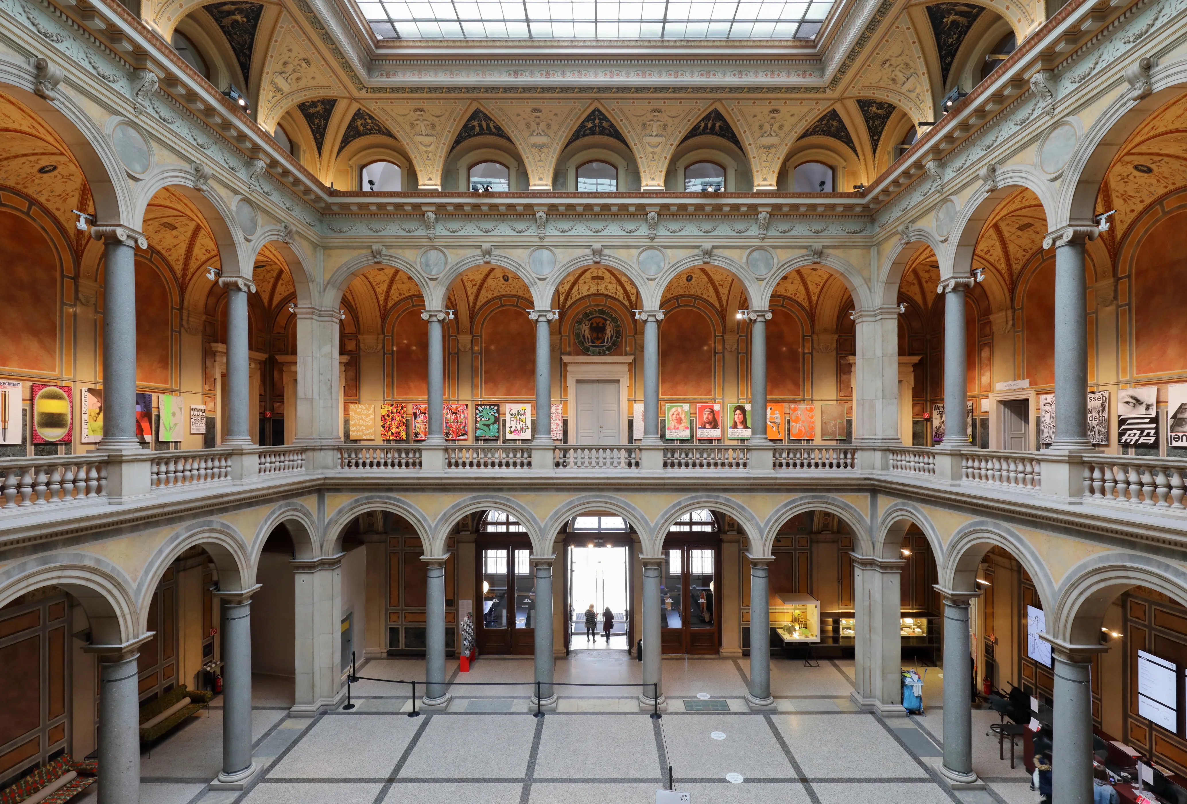 Een breed interieurbeeld van de Säulenhalle van het MAK Museum für angewandte Kunst, met gewelfde zuilengangen over meerdere verdiepingen, stenen zuilen en een glazen dak met tentoonstellingen langs de wanden.