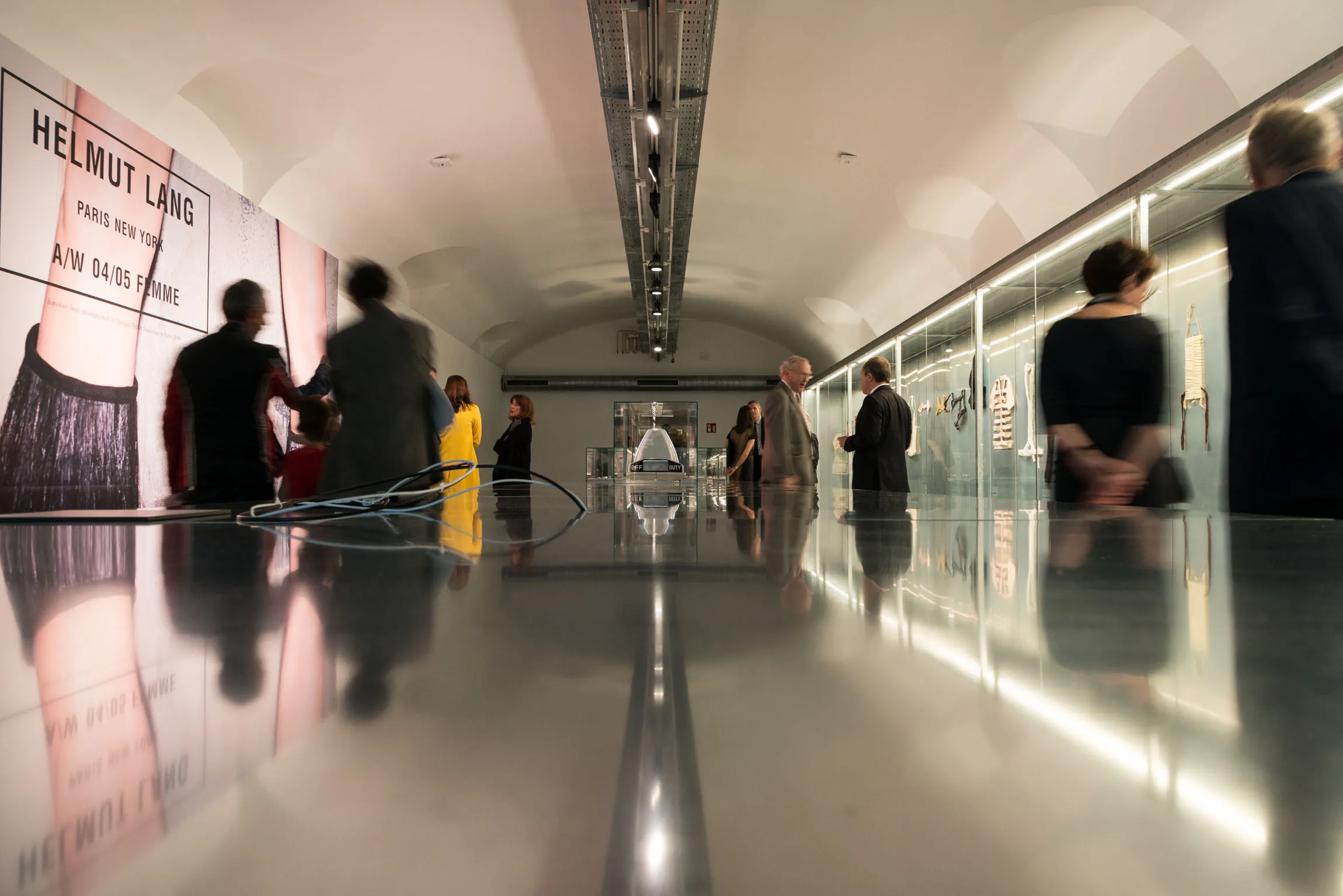 Tentoonstellingsruimte met een reflecterende tafel, mensen en een Helmut Lang-advertentie aan de muur