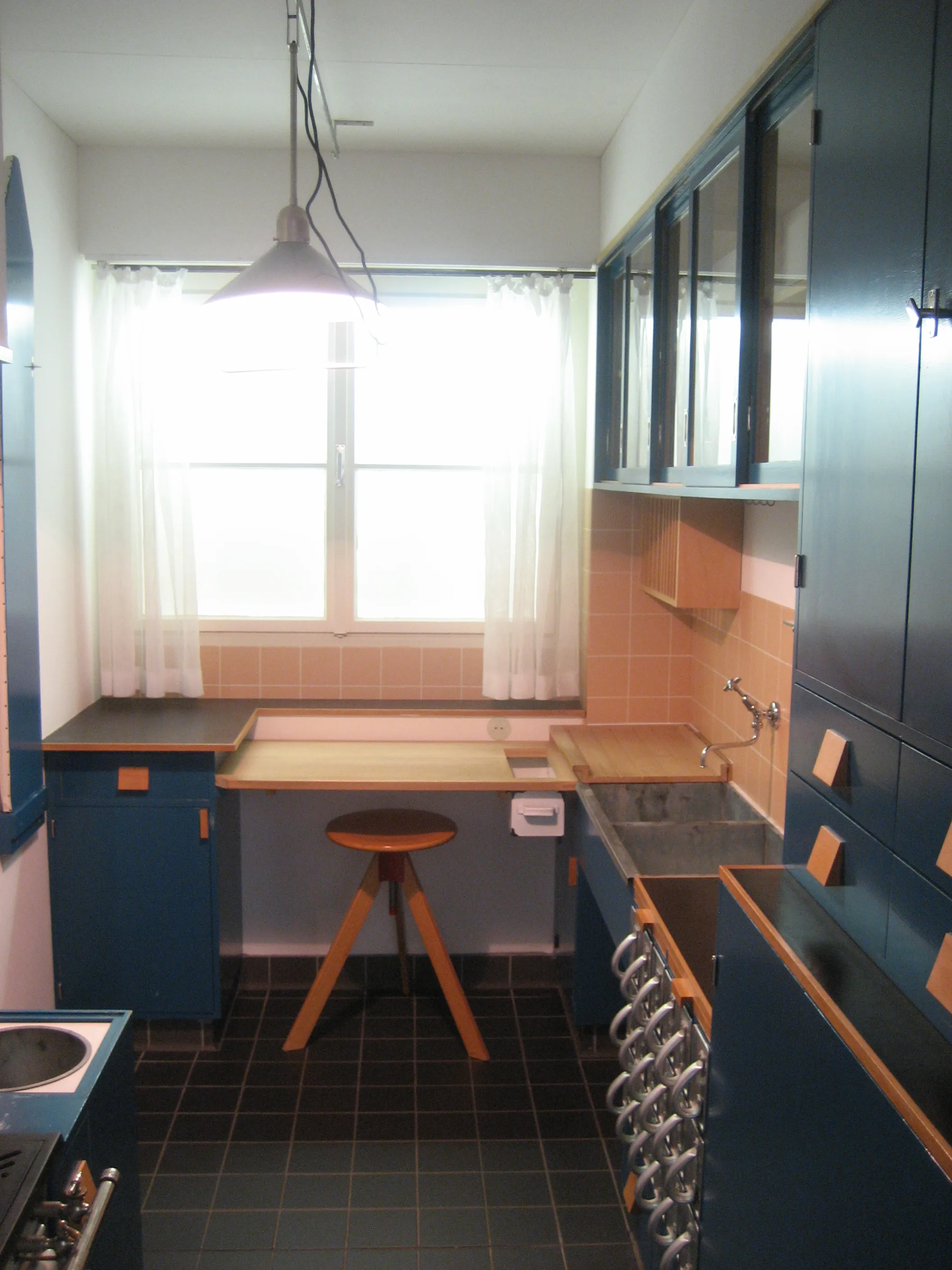 Interieur van een keuken met blauwe kasten, een houten kruk en een raam met vitrage
