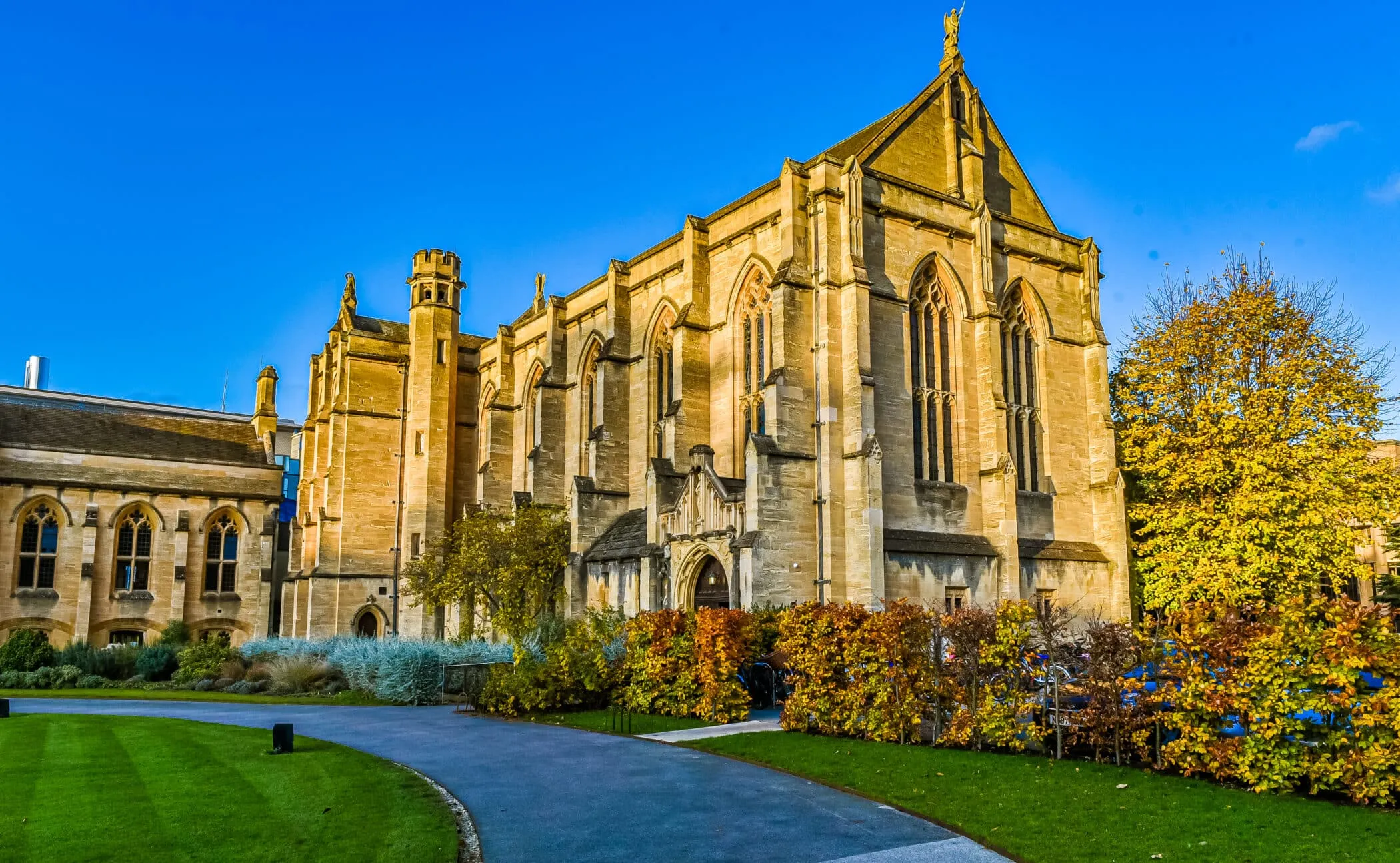 Capilla de piedra de estilo gótico del Mansfield College con follaje otoñal, camino pavimentado y césped verde bajo un cielo azul despejado