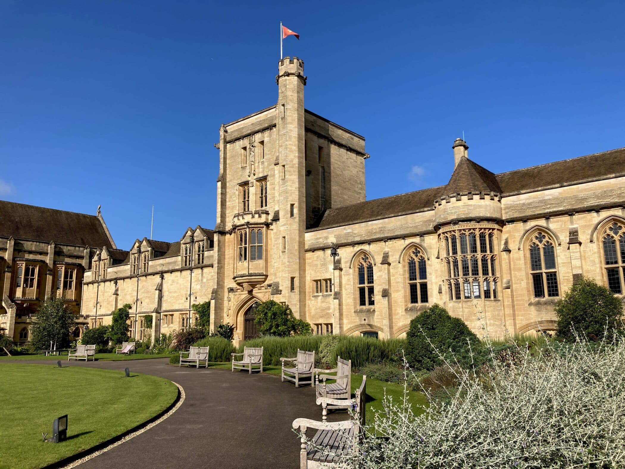 Fachada de piedra del Mansfield College con una torre central donde ondea una bandera, ventanas arqueadas, un césped bien cuidado y sillas de jardín de madera bajo un cielo azul despejado.