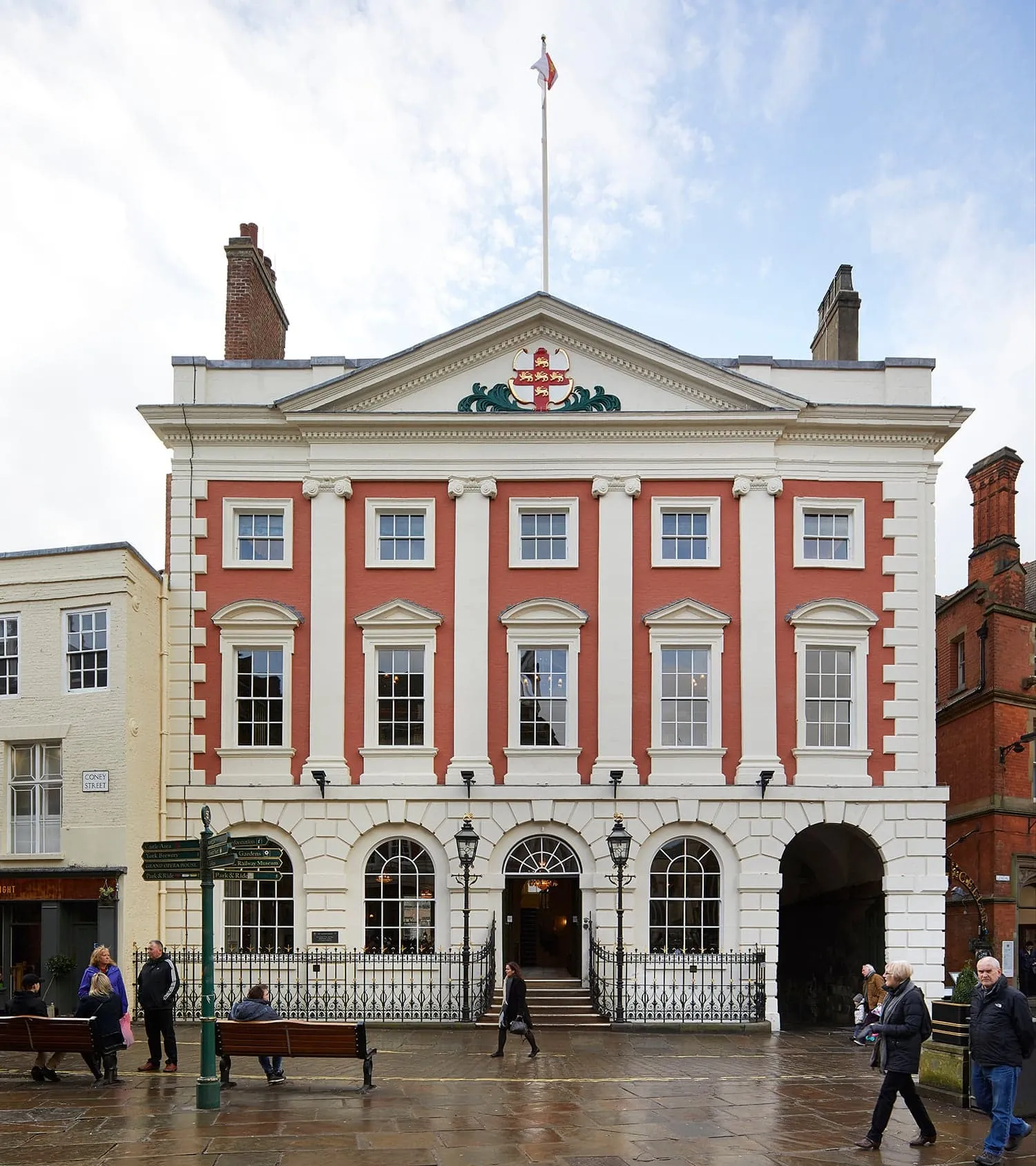 Georgiańska fasada York Mansion House z czerwonej cegły i białego kamienia, z łukowymi oknami na parterze, frontonem z herbem Yorku oraz masztem flagowym, z ludźmi spacerującymi po wybrukowanym placu przed budynkiem.