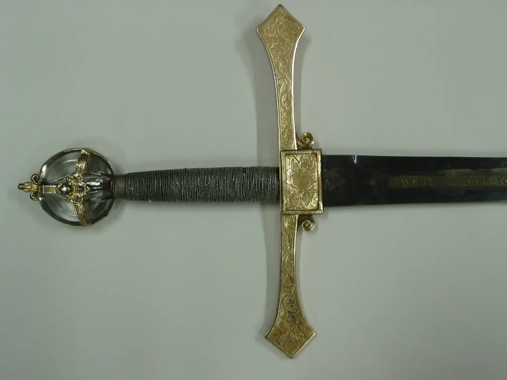 Miecz ceremonial z ozdobną złotą rękojeścią i jelcem, ciemnym owiniętym chwytem oraz okrągłą głowicą, zaprezentowany na prostym, jasnym tle