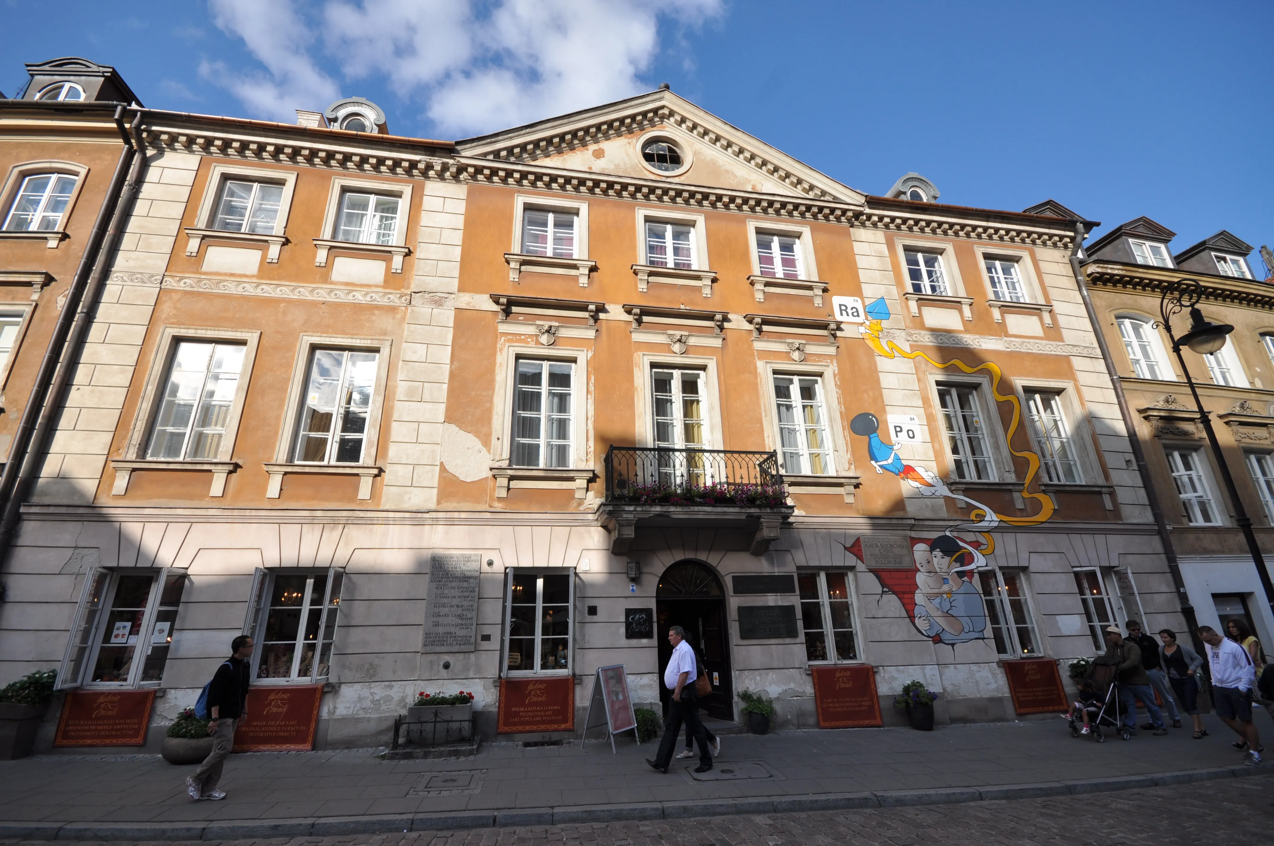 Klasycystyczny budynek z pomarańczowo-beżową fasadą, wieloma oknami i centralnym wejściem pod trójkątnym frontonem, z kolorowym muralem na ścianie bocznej i ludźmi spacerującymi po chodniku