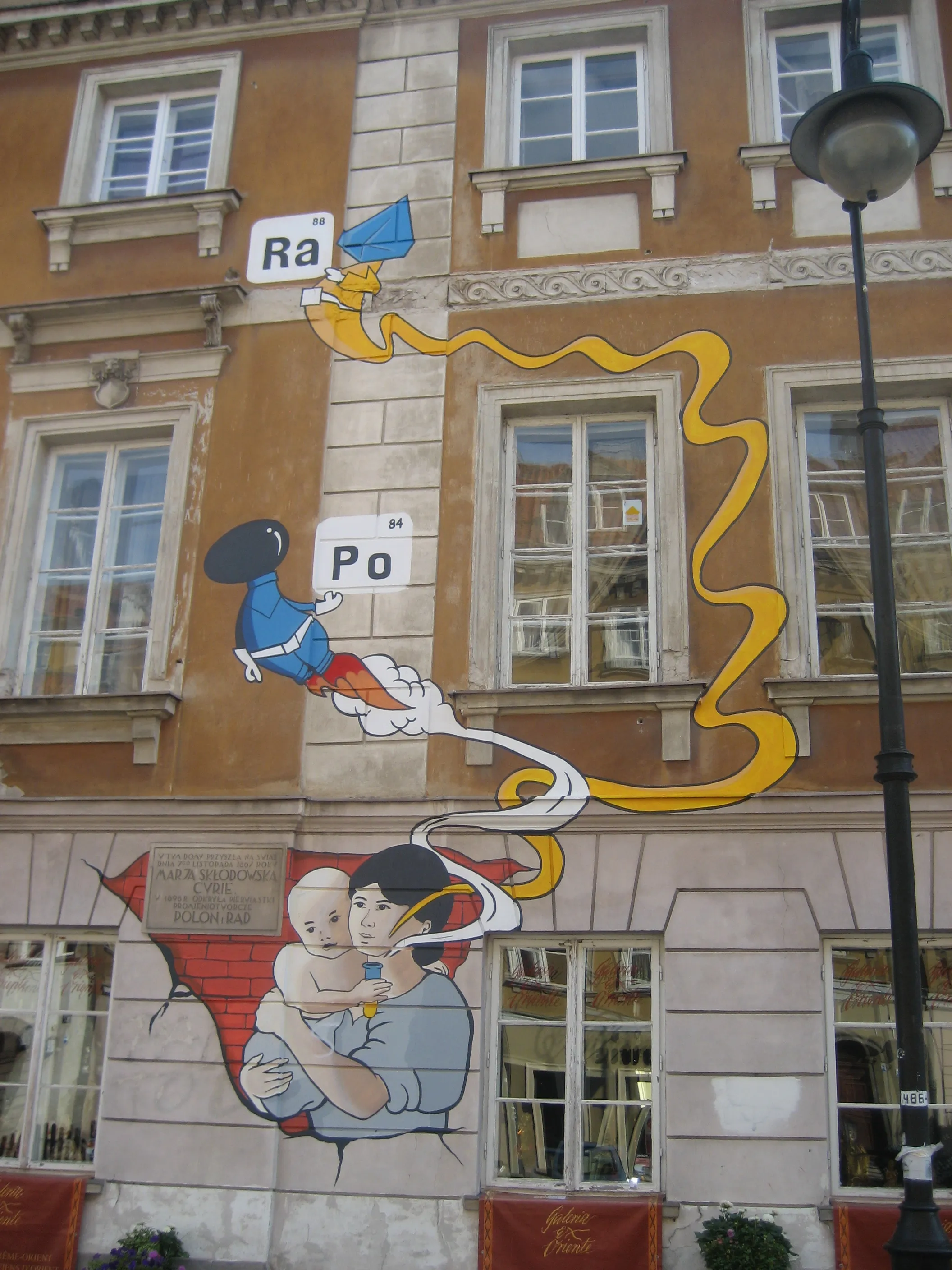 Mural na fasadzie budynku przedstawiający kreskówkowego naukowca z symbolami atomowymi i kobietę trzymającą dziecko.
