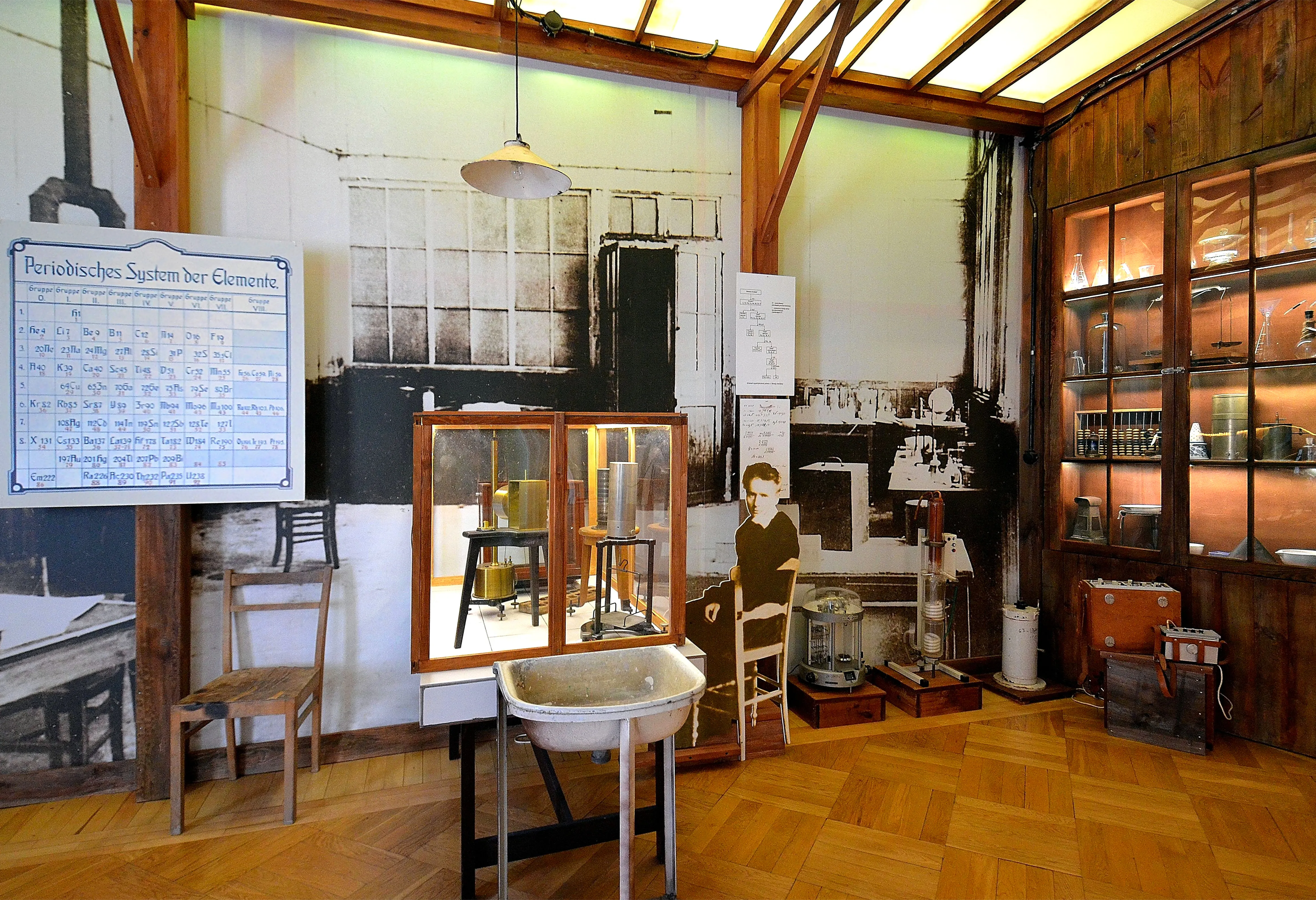 Wnętrze muzeum z drewnianymi ścianami i podłogami, gablotami, sprzętem naukowym i dużym panelem informacyjnym na ścianie.