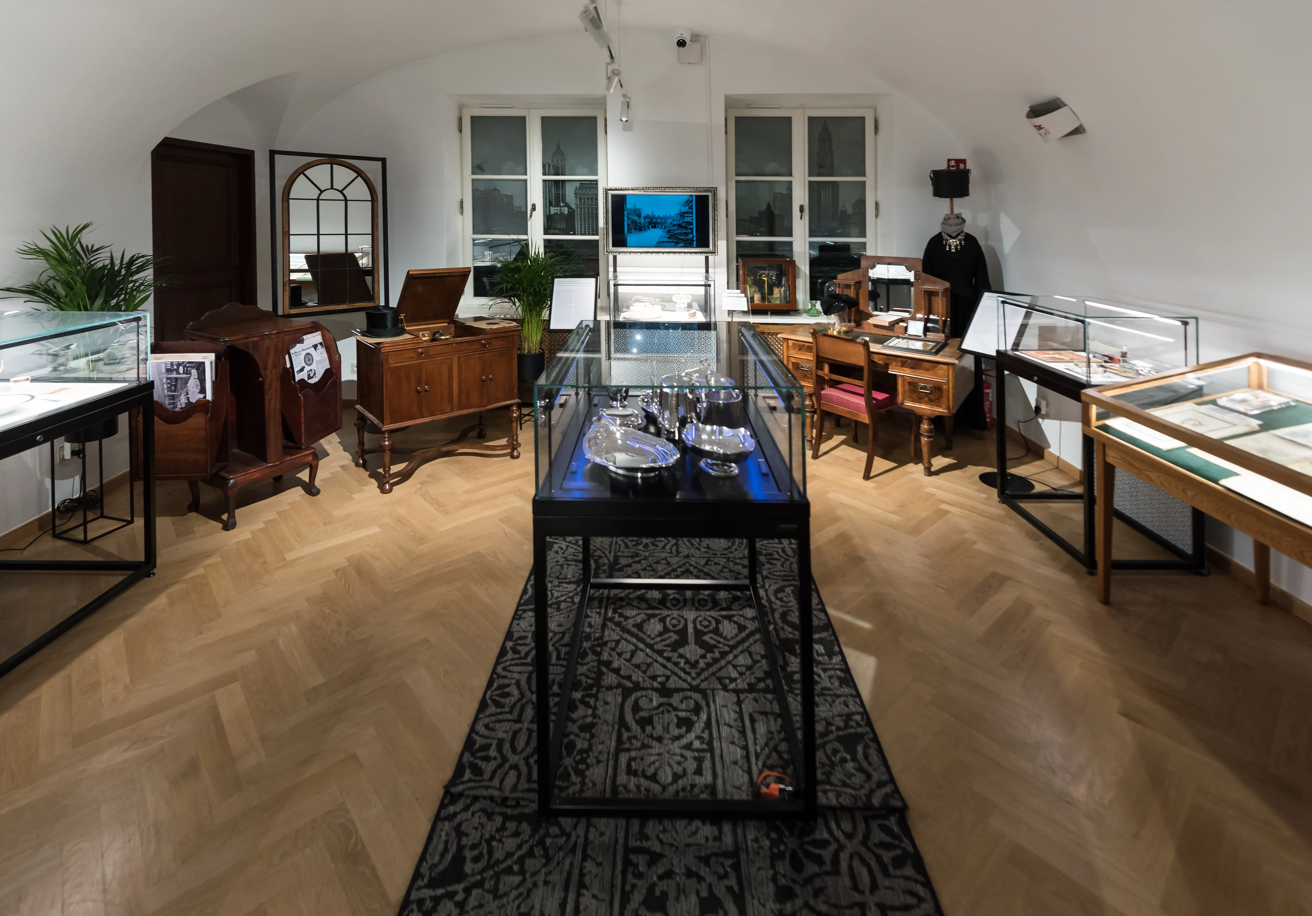 Wnętrze muzeum z gablotami, drewnianymi meblami i wzorzystym dywanem na podłodze w jodełkę