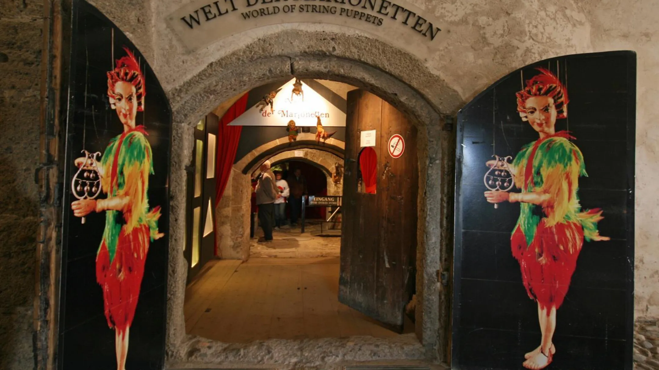 Gewelfde stenen ingang van het World of String Puppets museum, geflankeerd door twee grote beschilderde panelen met kleurrijke marionettenfiguren die klokken vasthouden