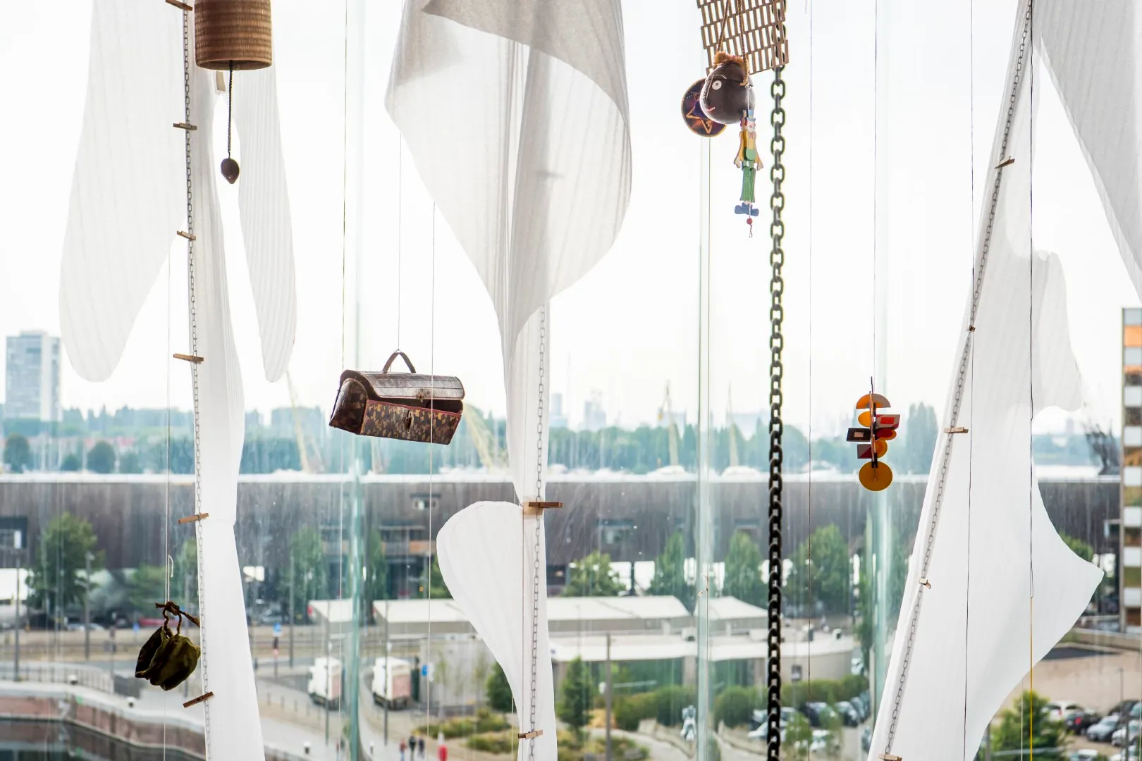 Instalación artística colgante con paneles de tela blanca, un bolso, una cesta y objetos de madera suspendidos por cadenas frente a grandes ventanales con vistas al paisaje urbano de Amberes.
