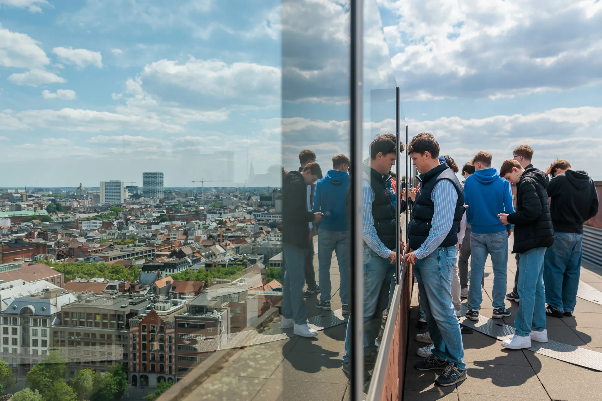 Grupo de visitantes de pie junto a la barandilla de cristal de una terraza en la azotea con vistas al paisaje urbano de Amberes bajo un cielo parcialmente nublado