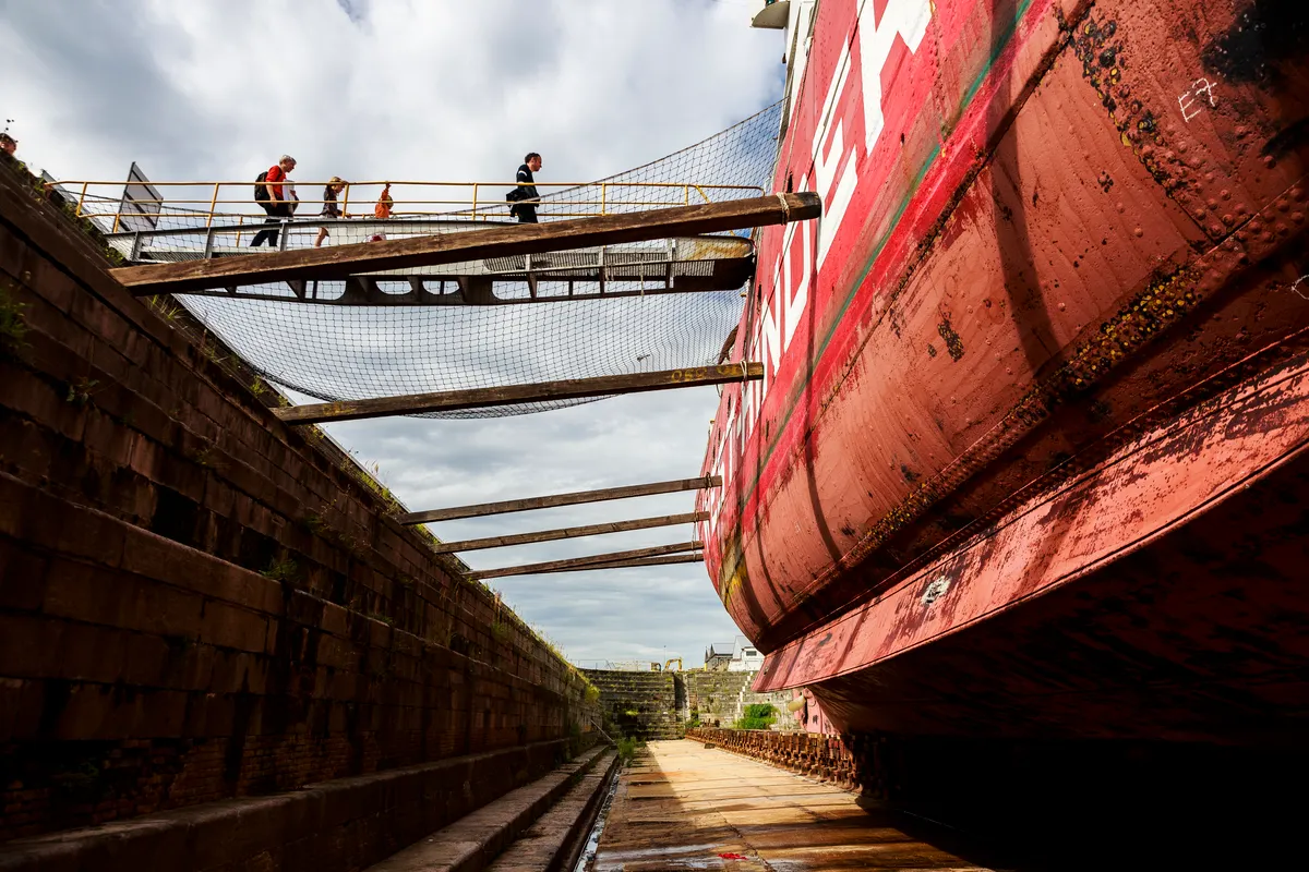 Gran casco de barco rojo descansando en un dique seco de piedra histórico con visitantes caminando por una pasarela metálica elevada