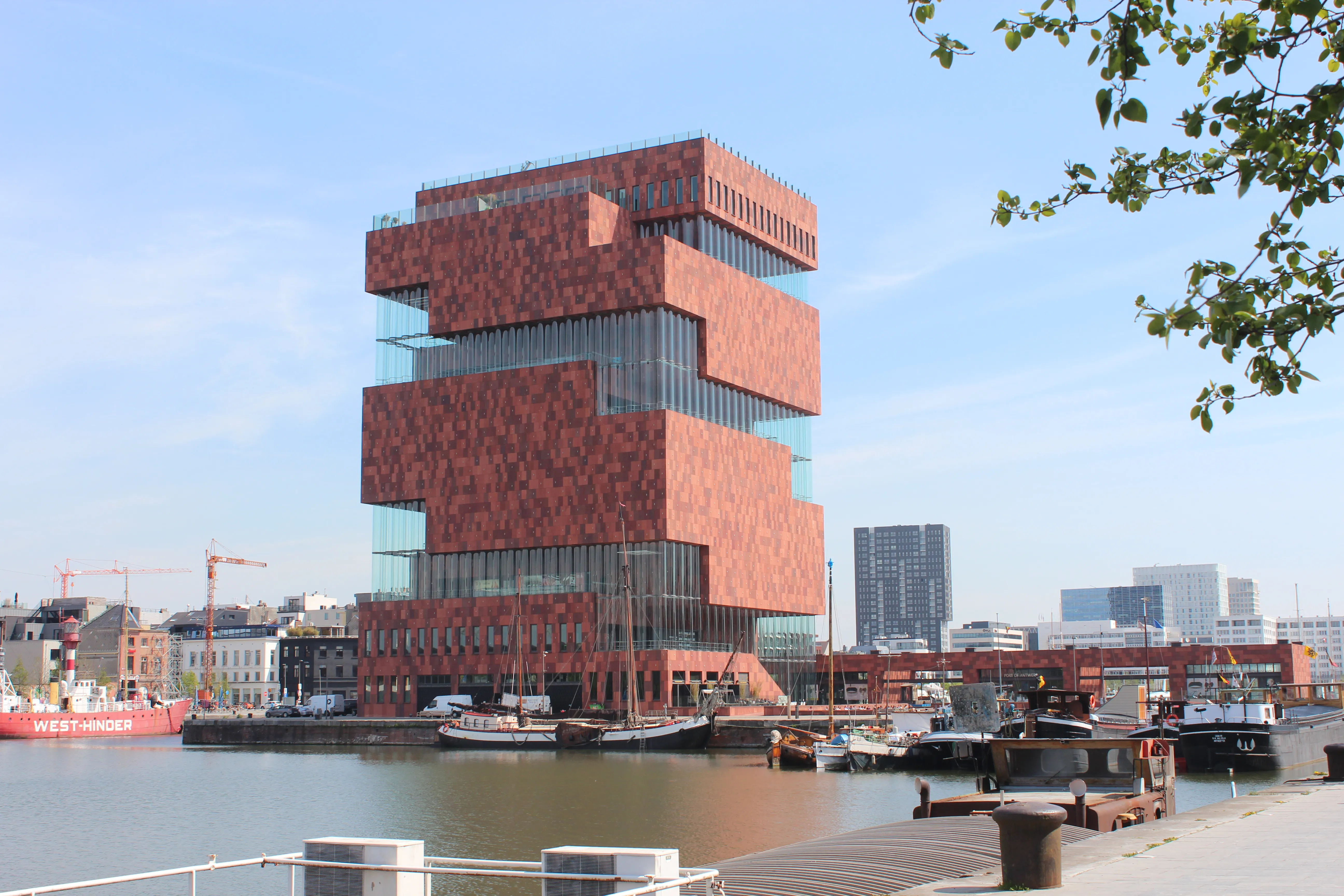 El edificio del museo MAS de arenisca roja, con volúmenes en forma de cajas apiladas y desplazadas que se elevan sobre el paseo marítimo de Amberes, con barcos amarrados a lo largo del muelle y un cielo azul despejado.