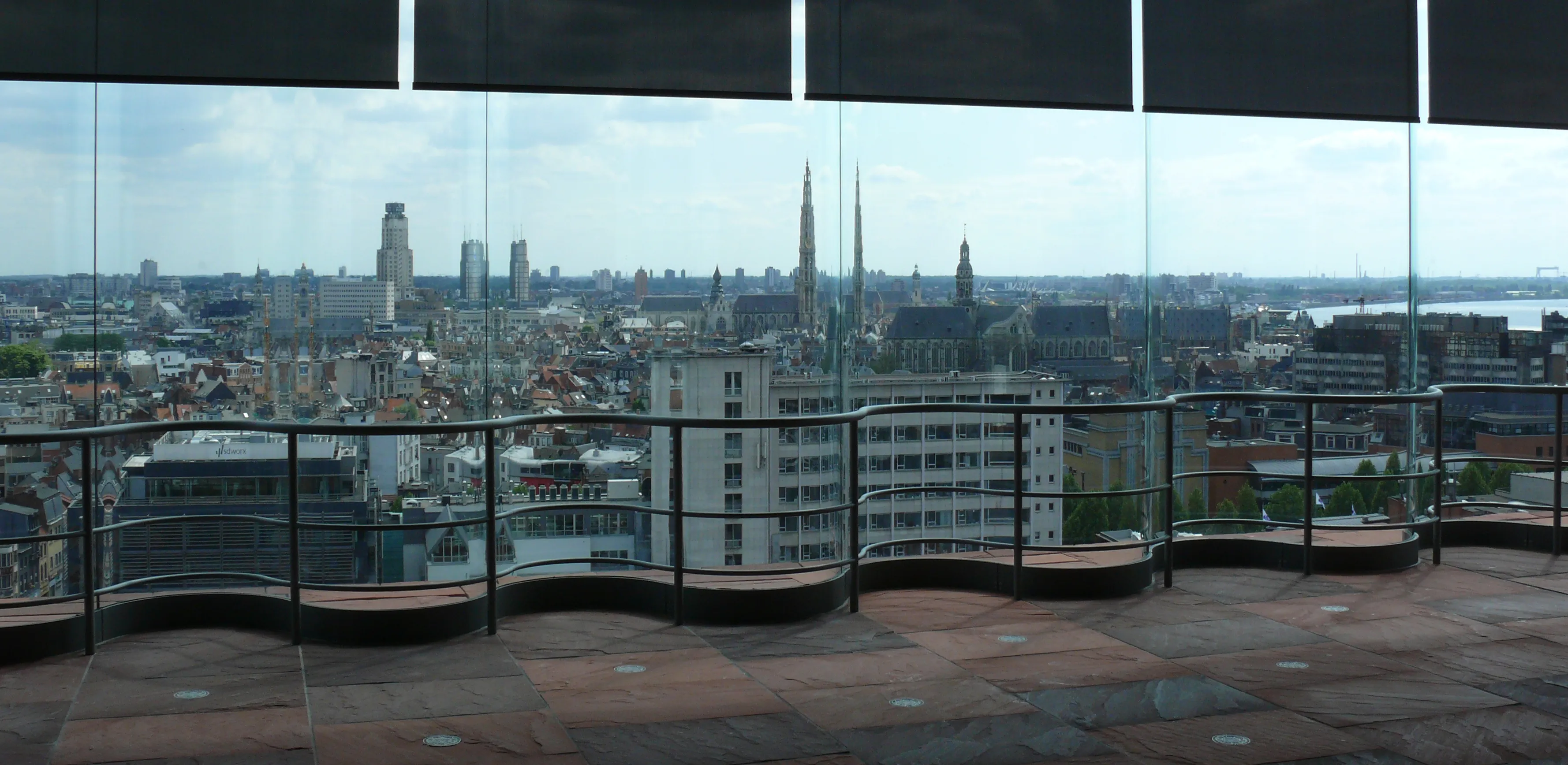 Vista panorámica del paisaje urbano de Amberes con torres de iglesias y edificios vistos a través de paneles de cristal desde la terraza de la azotea del MAS, con bancos curvos.