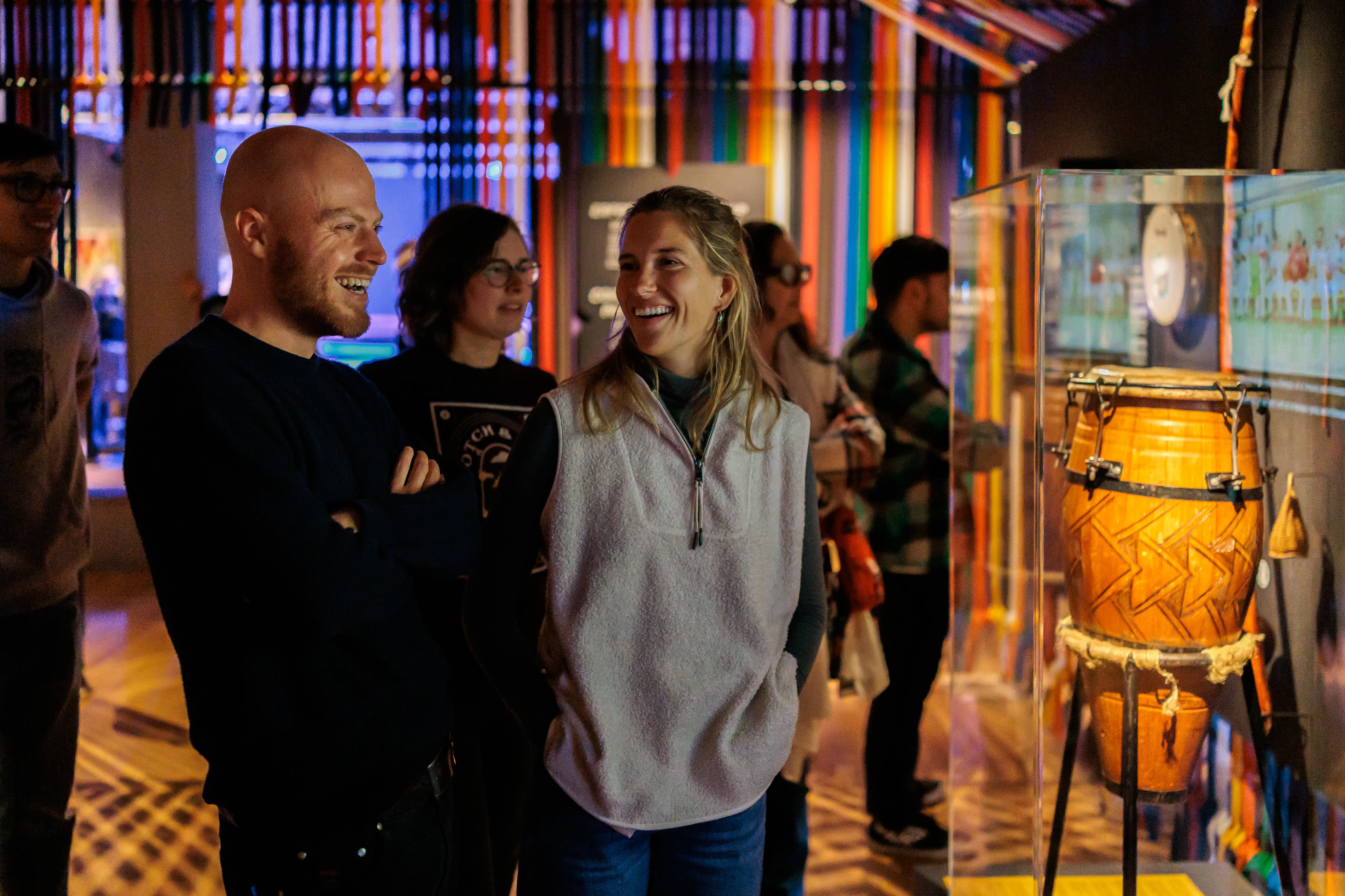 Visitantes sonriendo mientras observan un tambor decorado en una vitrina de cristal en el museo MAS, con coloridas tiras verticales colgando al fondo