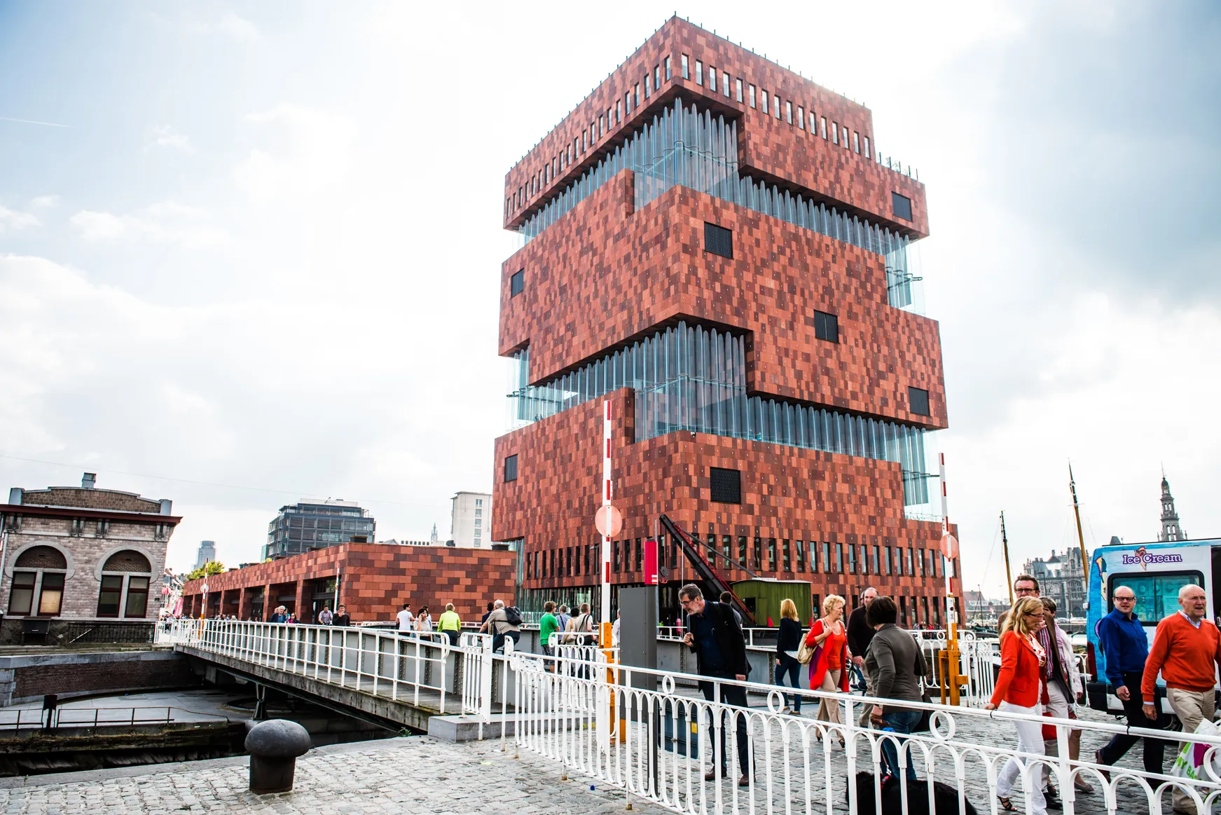 El edificio del MAS Museum aan de Stroom en Amberes, una alta estructura de ladrillo rojo con volúmenes superpuestos desplazados y fachadas de cristal, visto desde el paseo marítimo con gente caminando por un puente con barandilla blanca.