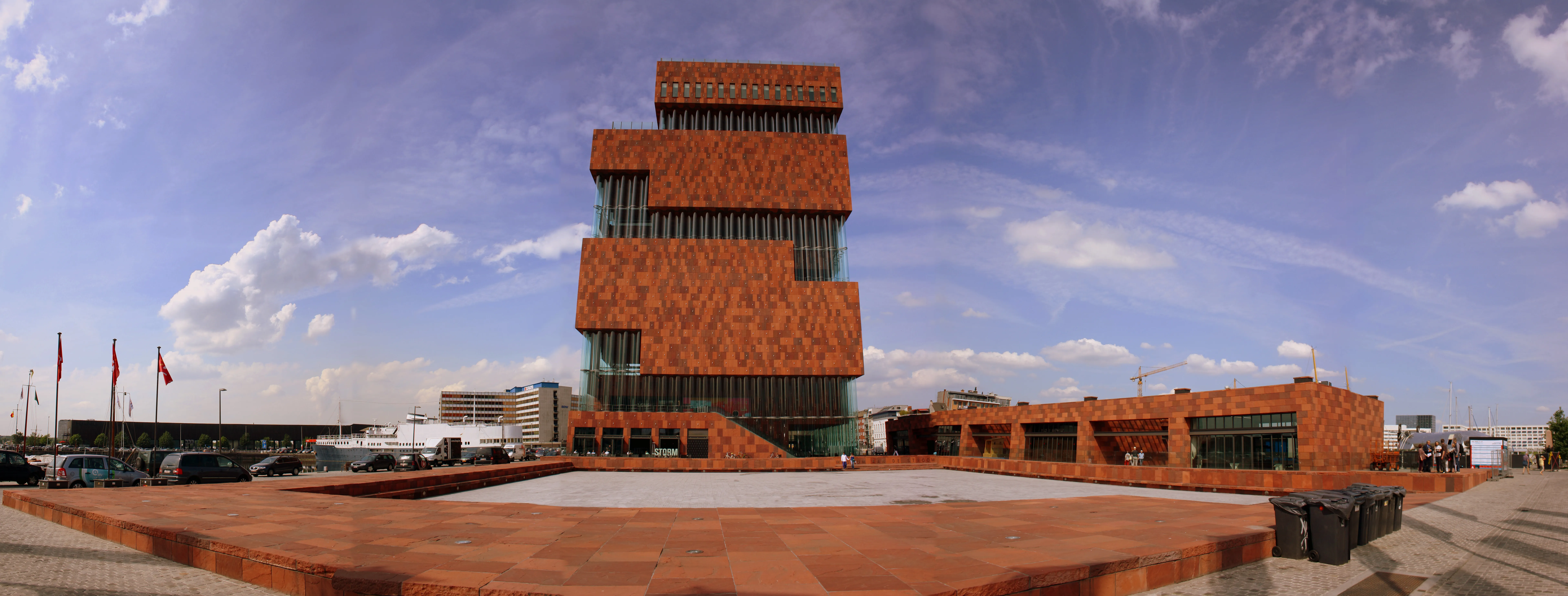 Vista panorámica del edificio de ladrillo rojo del Museum aan de Stroom, que presenta una arquitectura de cubos apilados y una gran plaza pavimentada bajo un cielo azul.