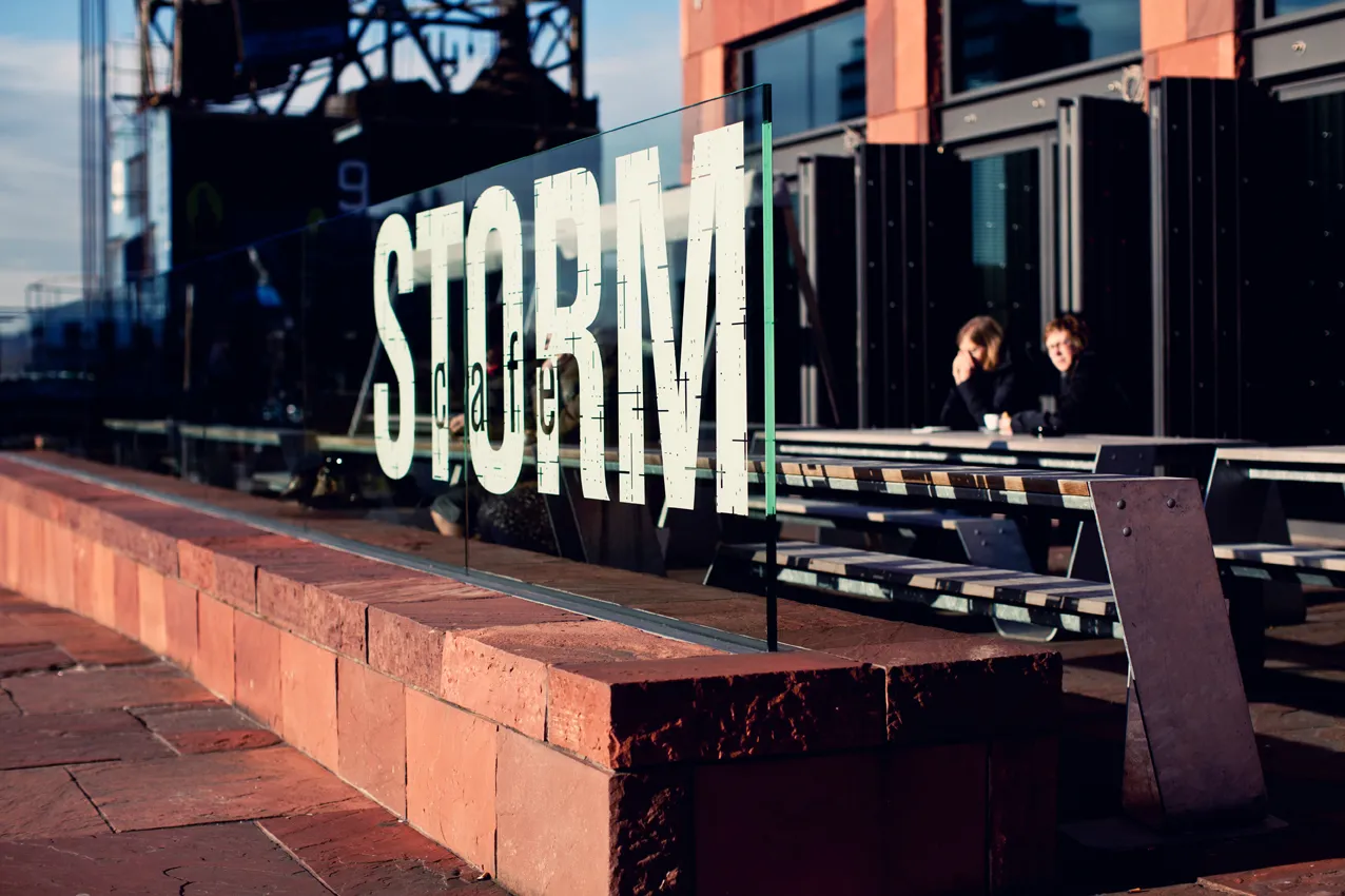 Terraza exterior en la azotea con letras blancas grandes de STORM en una barrera de cristal, paredes de arenisca roja y personas sentadas en mesas al fondo.