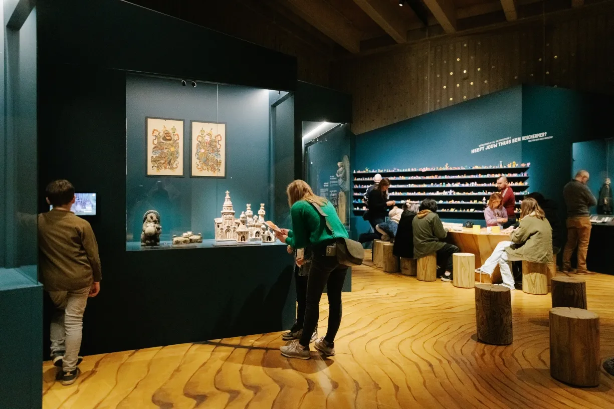 Interior de museo con visitantes observando una maqueta arquitectónica y grabados enmarcados expuestos sobre paredes color cerceta, con taburetes de tronco de madera dispuestos sobre un suelo de madera con patrón ondulado.