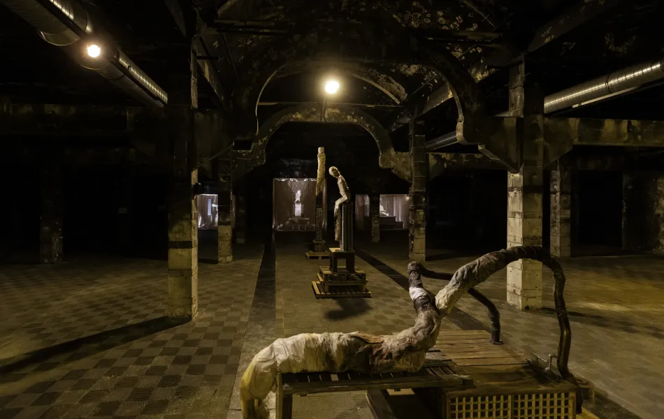 Dark interior space with industrial architecture featuring arched ceilings and exposed pipes, displaying contemporary art installations including a large sculpture and a tall figure.