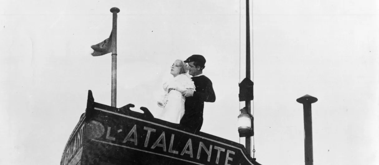 A black-and-white photo of a man and woman embracing on a ship's bow with the text 'L'ATALANTE' on the hull