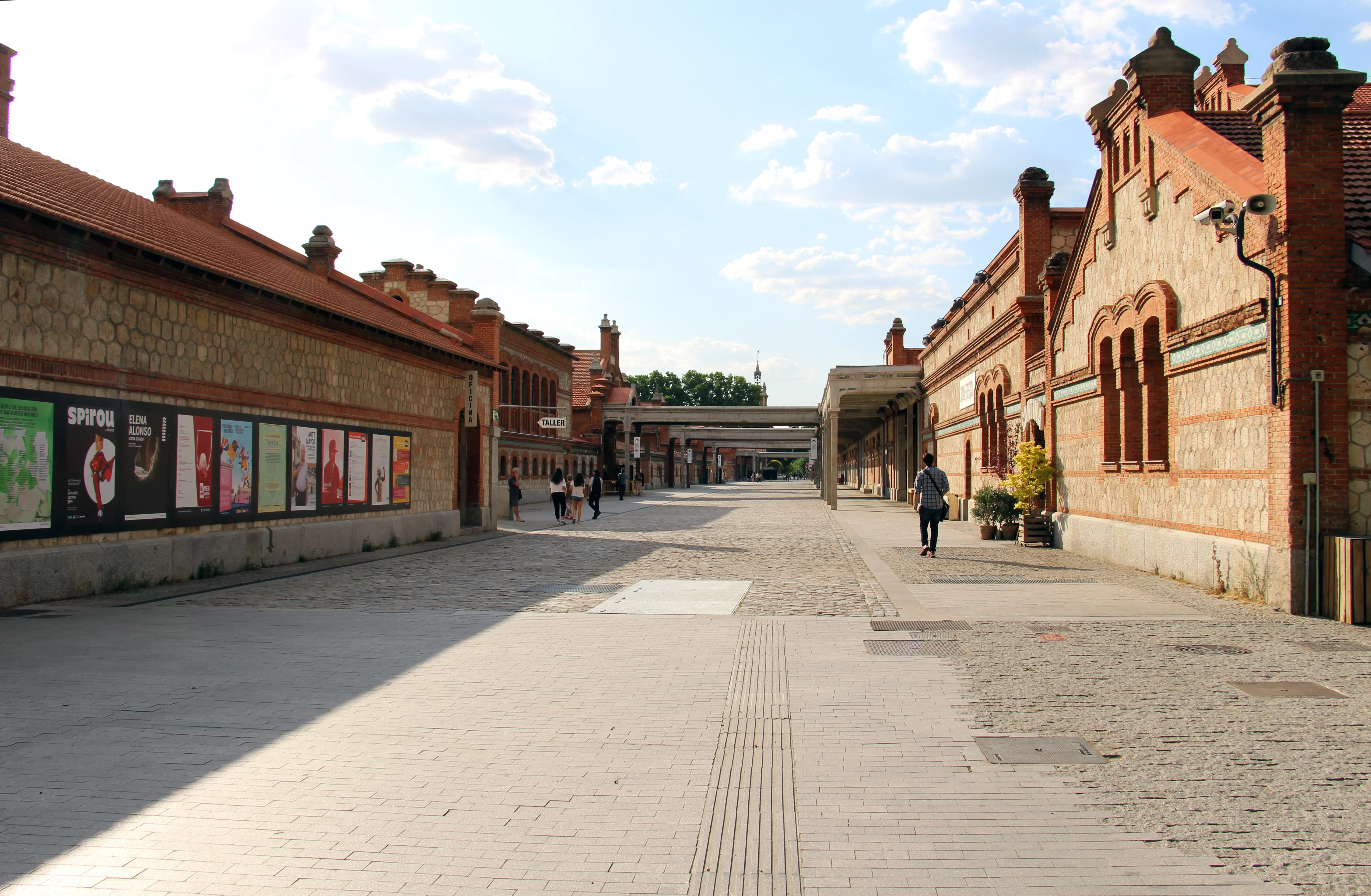 Obszerny dziedziniec między dwoma ceglanymi budynkami przemysłowymi w Matadero Madrid, z widoczną w oddali osobą spacerującą.