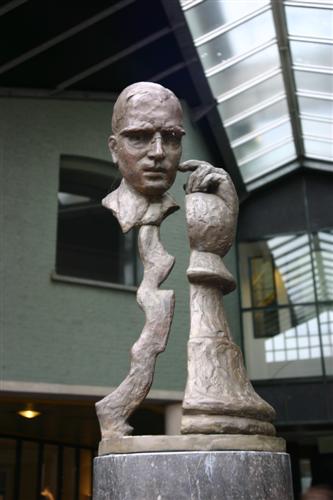 Bronze bust sculpture of a man integrated with a chess king piece, displayed inside a museum with glass ceiling and modern interior architecture
