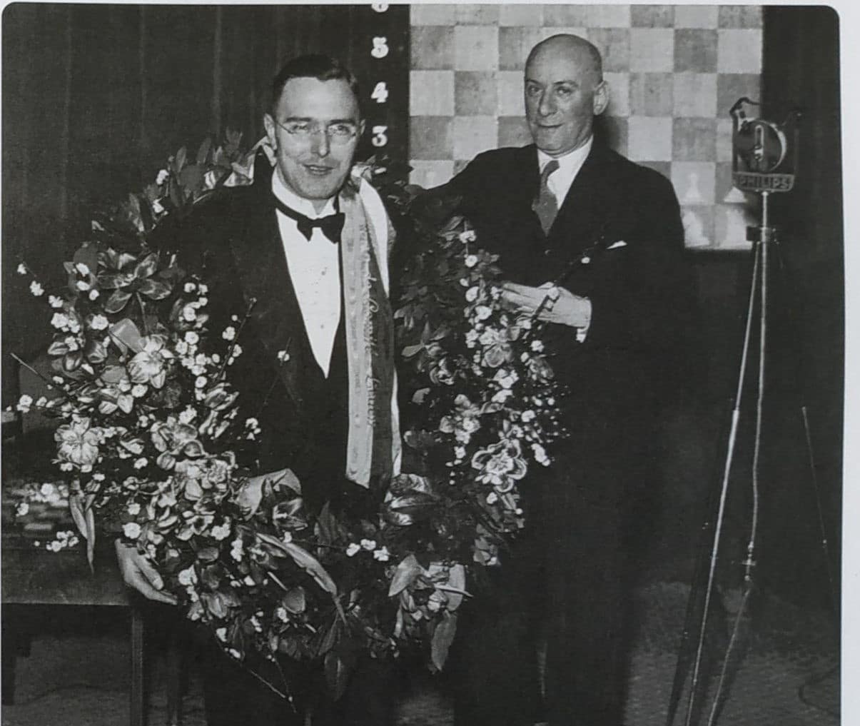 Two men in formal attire holding large floral arrangements with a checkered wall and vintage microphone in the background