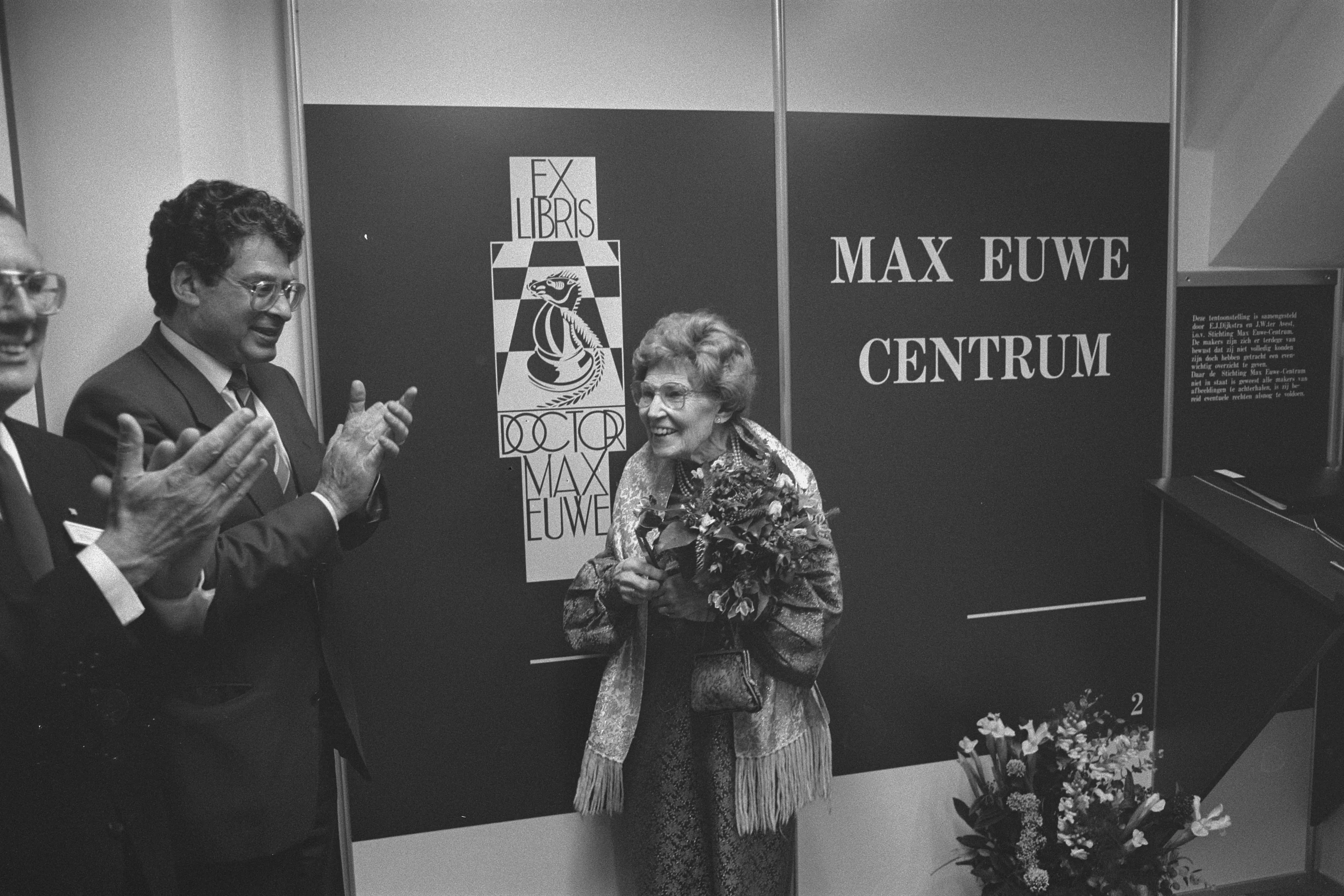 Two men clapping and a woman holding flowers in front of a sign that reads 'MAX EUWE CENTRUM' with a chess-themed emblem.