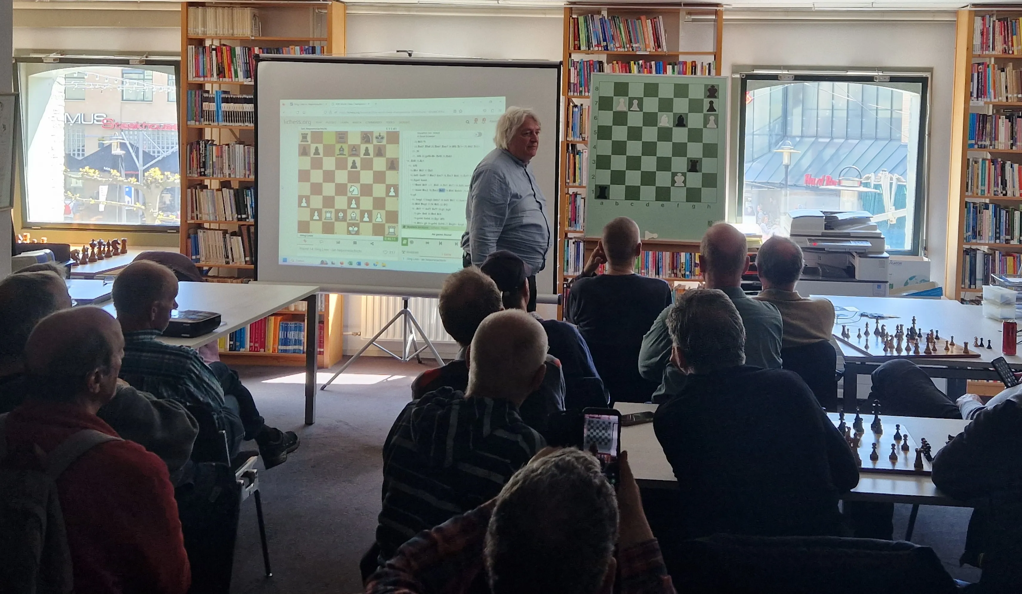 A man standing in front of a projection screen showing a chessboard, addressing a seated audience in a room with bookshelves and chessboards.