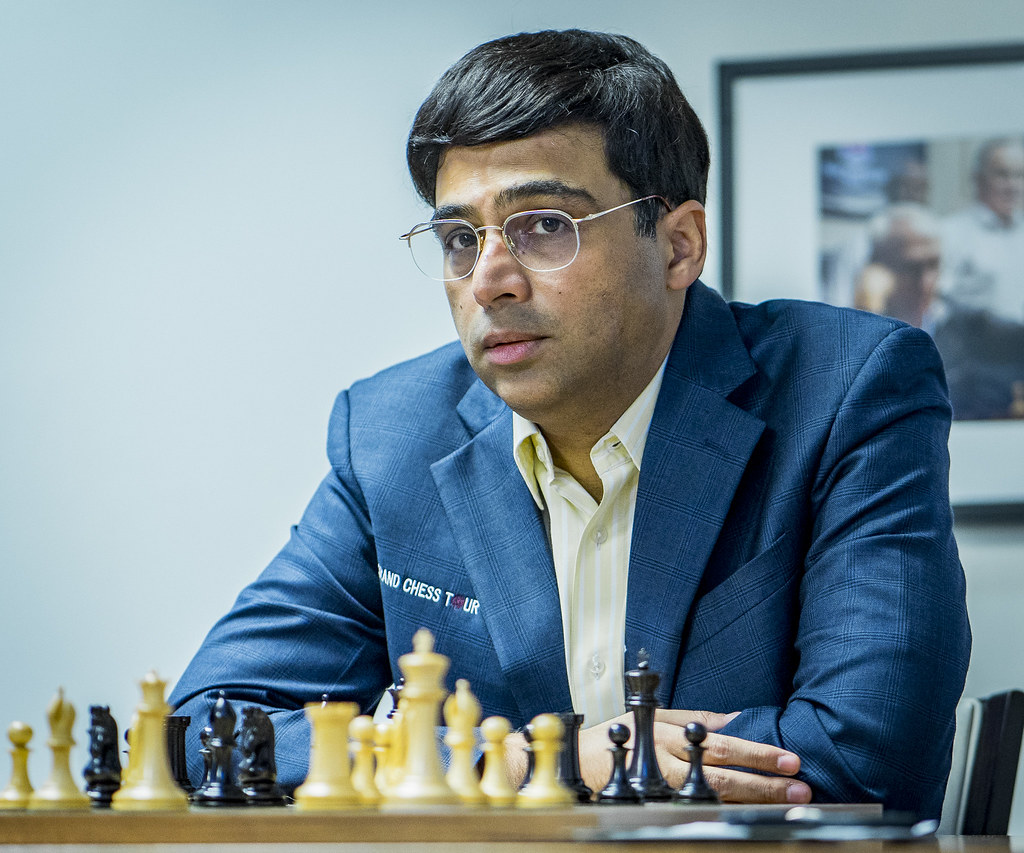 Person in blue suit seated at chessboard with black and white pieces, indoor setting with framed photos on wall