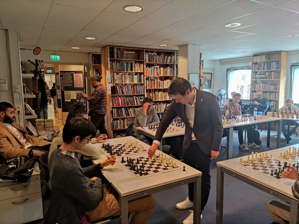 A man in a dark jacket playing chess at a table surrounded by other people playing chess in a room with bookshelves