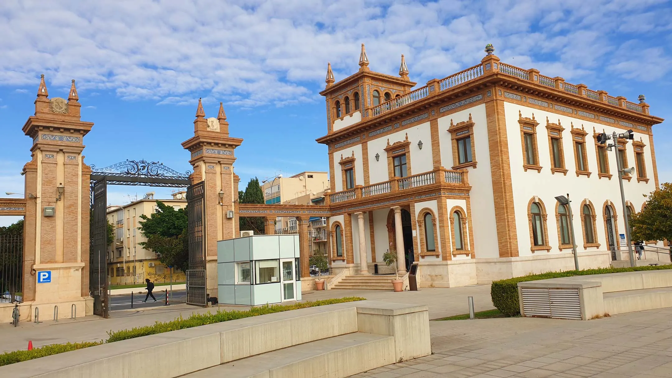 Vista exterior del histórico complejo de la Tabacalera en Málaga, mostrando la ornamentada puerta de entrada con sus torres gemelas y el edificio principal blanco con detalles en terracota bajo un cielo azul