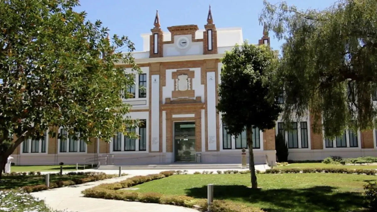 Exterior de un edificio histórico de ladrillo y blanco con ventanas y entrada arqueadas, rodeado de árboles verdes y un césped bajo un cielo azul.