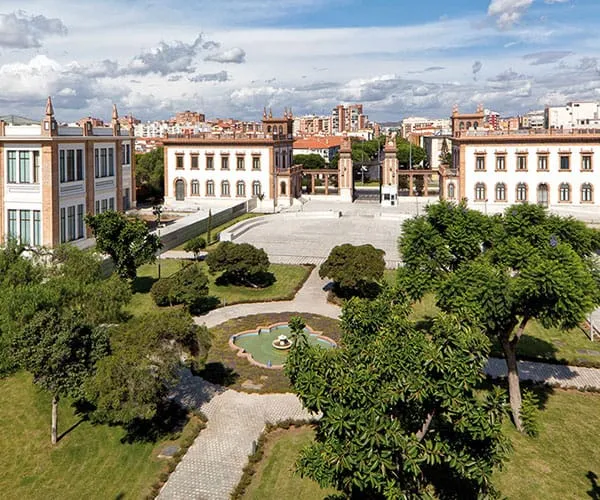 Vista elevada del patio del histórico complejo de la Tabacalera con sus edificios de ladrillo circundantes, una plaza pavimentada, senderos ajardinados, árboles y una pequeña fuente