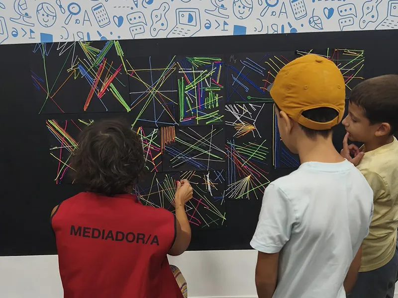 Tres niños están frente a una pizarra grande cubierta de dibujos hechos con tiza de colores; uno de ellos lleva un chaleco rojo con la etiqueta MEDIADOR/A mientras los otros observan la instalación de arte interactiva.