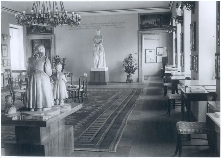 Interior of a museum gallery with a long carpet, wooden tables and chairs, and statues including a white marble figure of a woman in the center.