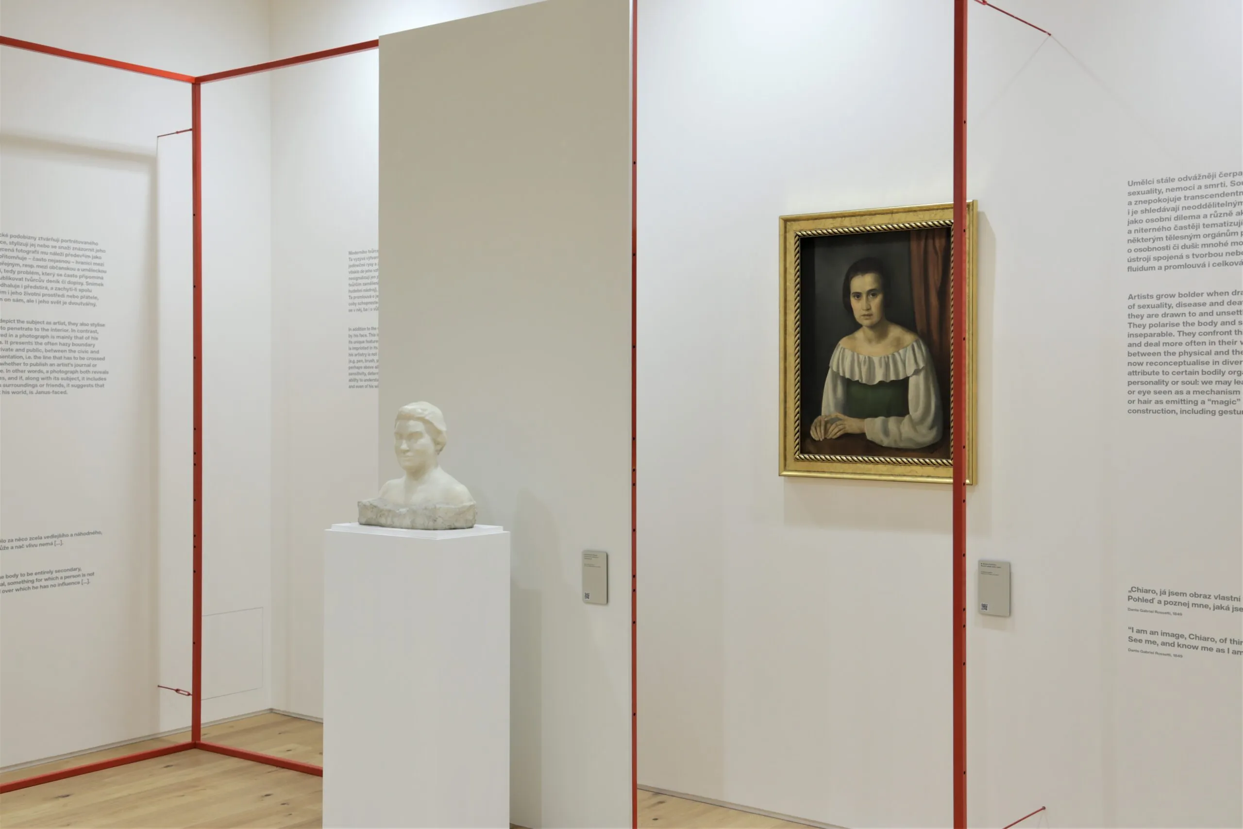 Een museumgalerij met een ingelijst portretschilderij, een wit marmeren buste op een sokkel en rode scheidingswanden op witte muren.