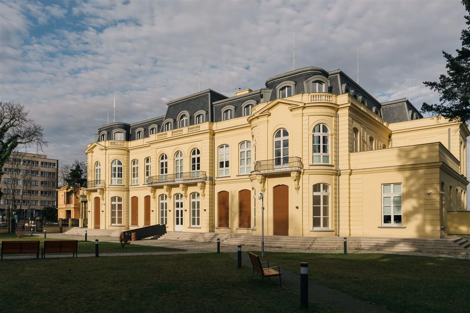 geel gebouw met boogramen en een mansardedak onder een bewolkte hemel