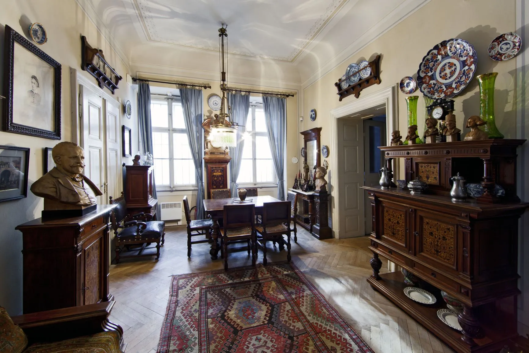 Interieur van een historische kamer met houten meubels, een buste, ingelijste portretten en decoratieve borden aan de muren en een kast.