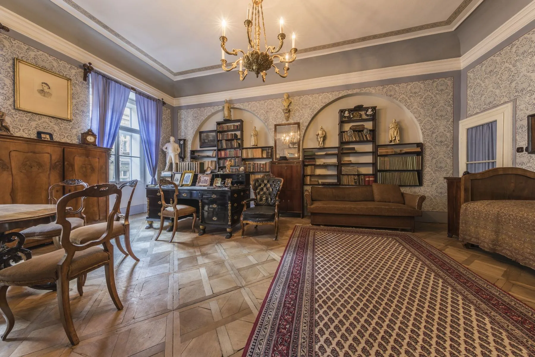 Een historische kamer met houten meubels, boekenplanken, een kroonluchter en een behang met patroon.
