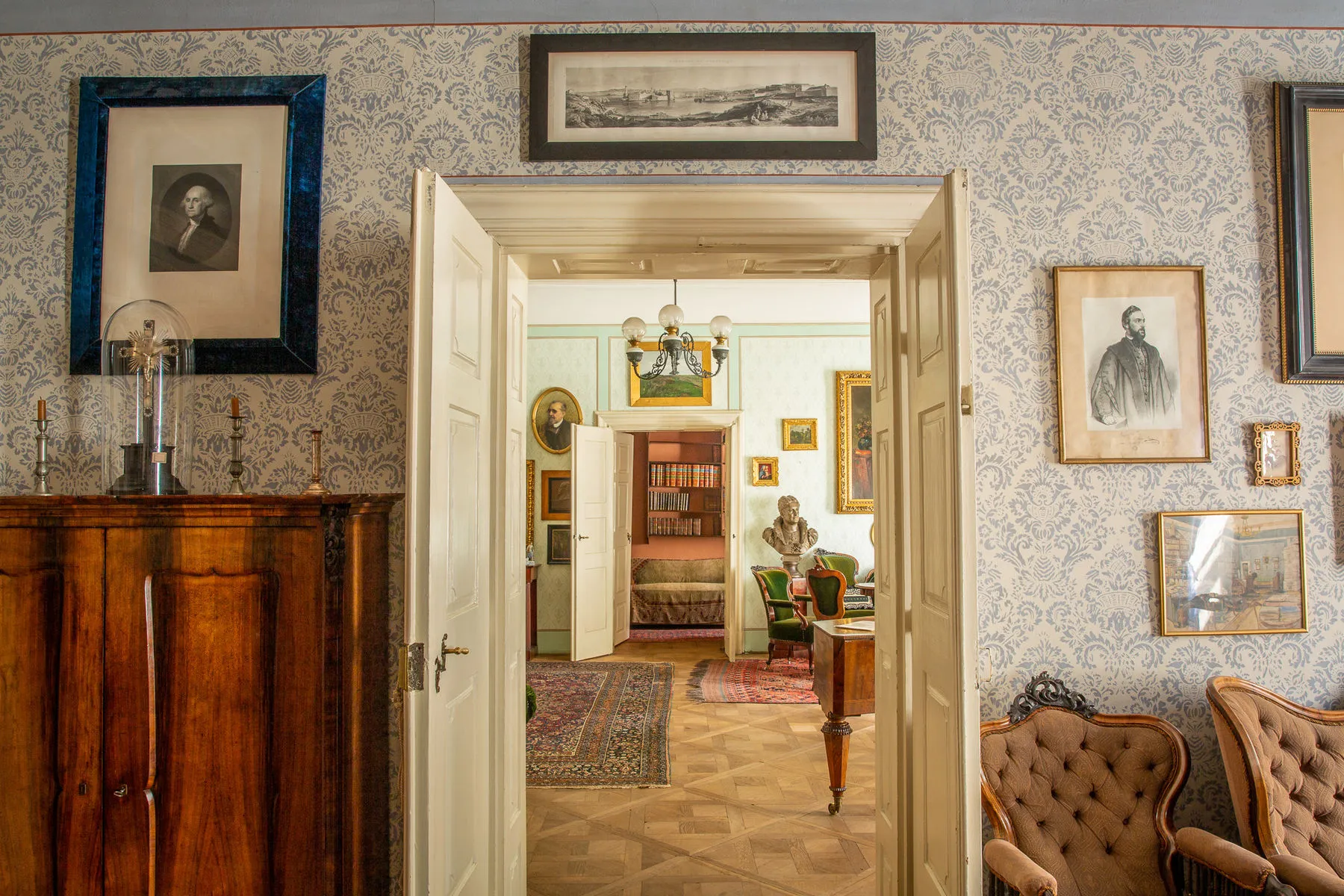 Een bewaard gebleven 19e-eeuwse kamer met patroonbehang, ingelijste portretten, houten meubels en uitzicht op een aangrenzende ruimte met een boekenkast en een borstbeeld.