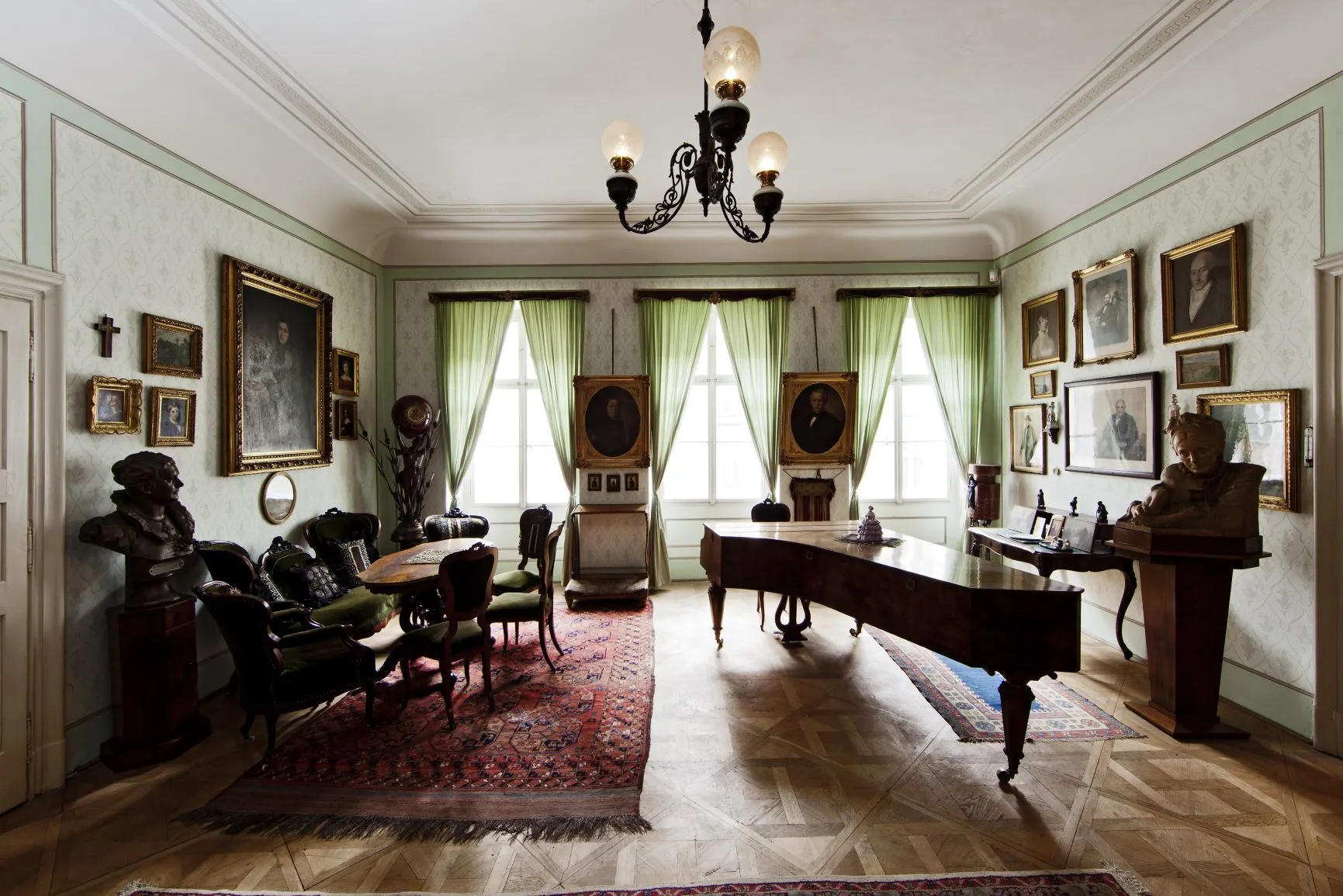 Een historische kamer met houten vloeren, een vleugel, een eettafel en ingelijste portretten aan lichtgroene muren.