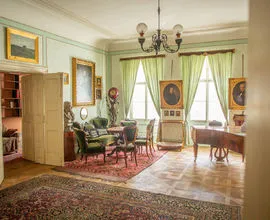 Interieur van een historische kamer met groene muren, houten meubels, ingelijste portretten en een kroonluchter.